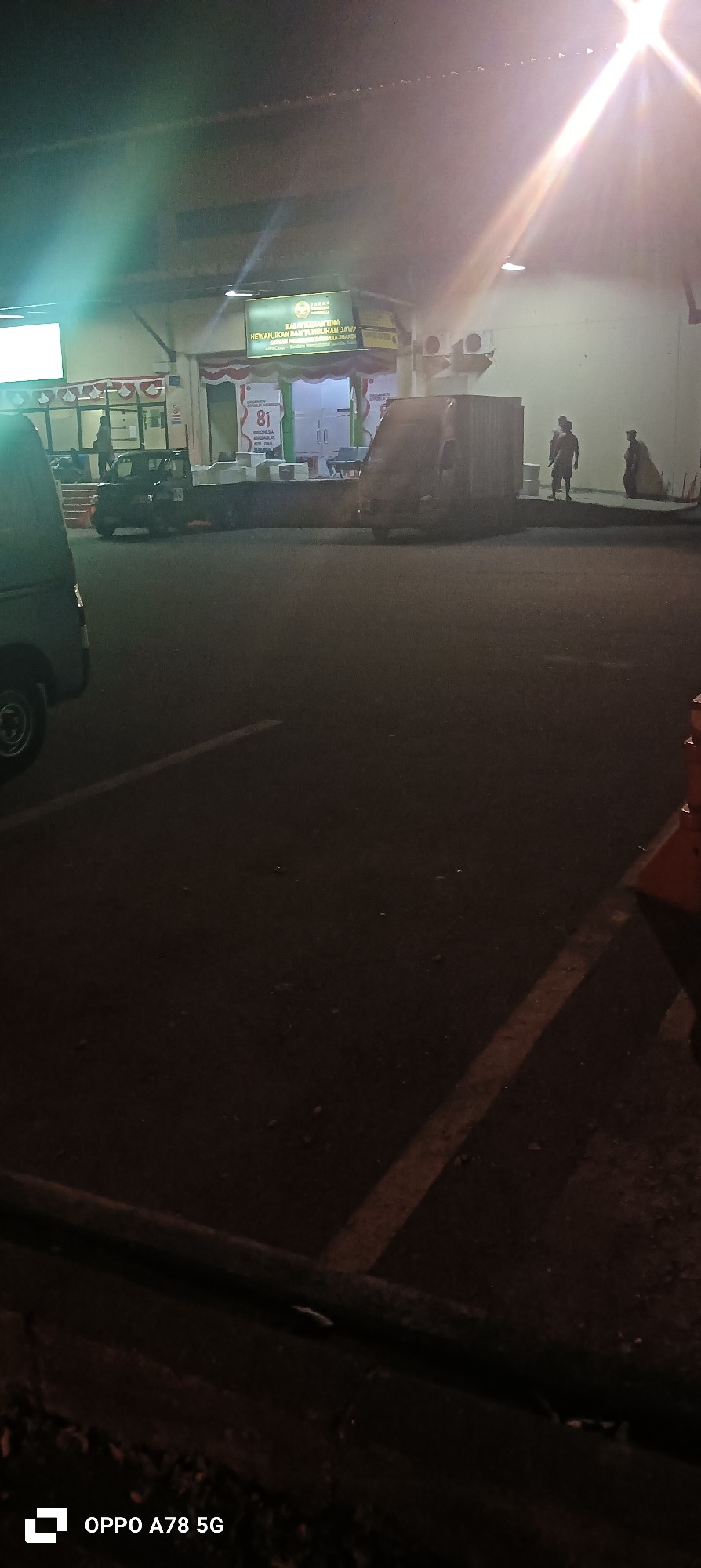 At night in the Semampir district of Indonesia, a building with an illuminated sign reads "BALAI KARANTINA HEWAN, IKAN DAN TUMBUHAN JAWA TIMUR". In front of the building, a small, dark-colored utility vehicle is parked to the left. A white box truck is positioned in the middle, with three individuals standing in front of it. To the right, another vehicle, possibly a van, is partially visible. The scene appears to be a loading or unloading area, with a bright light illuminating the area from the upper right corner of the frame. The ground is asphalt, with some debris scattered across it.