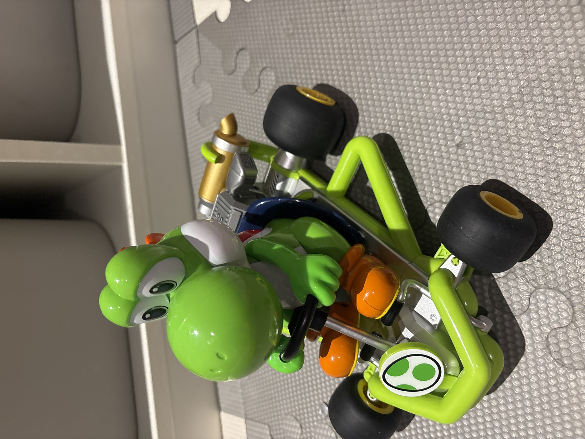 A green Yoshi toy character is seated in a green and silver toy go-kart. The go-kart has black wheels with yellow rims, and a white circular emblem with green polka dots on the front. Yoshi is wearing orange shoes and has large green eyes and a prominent snout. The toy is positioned on a grey textured surface. The background consists of light-colored shelving units. The scene does not provide any information to determine its location in Dubai, United Arab Emirates.