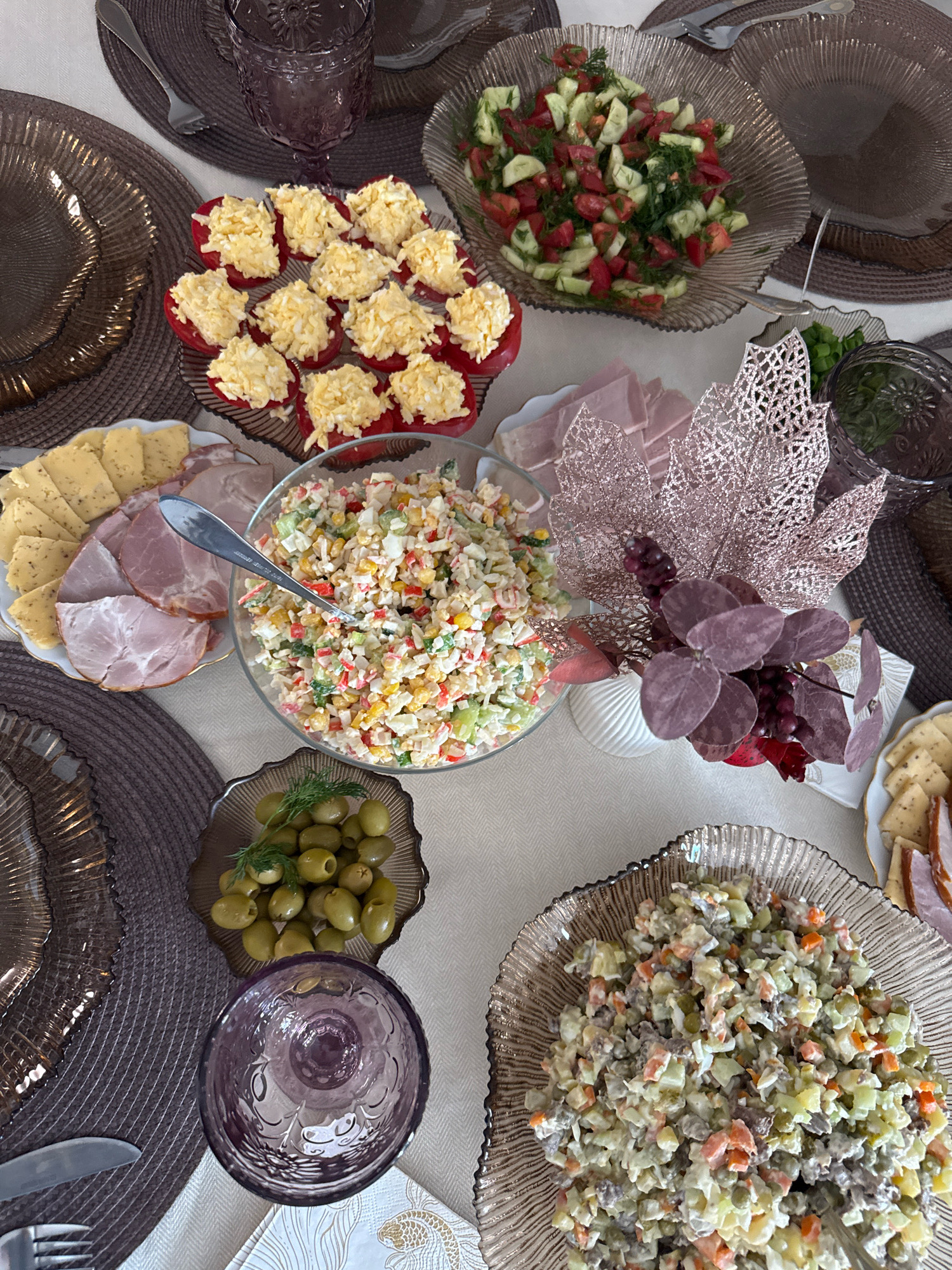 The image shows a table set for a meal, possibly in Saint Petersburg, Russia, though no specific landmarks or indicators of location are present. The table is covered with a light-colored tablecloth. Several dishes of food are arranged on the table, including a salad with tomatoes and cucumbers, stuffed red bell peppers topped with grated egg, a creamy salad with shredded vegetables and possibly crab sticks, a dish of green olives, a plate of sliced ham and cheese, and another salad composed of diced vegetables, meat, and a creamy dressing. There are also decorative elements on the table, including a vase with artificial flowers and metallic leaf decorations, and several ornate purple glasses. Place settings are visible, with chargers, plates, and cutlery. The overall impression is of a festive gathering or meal.