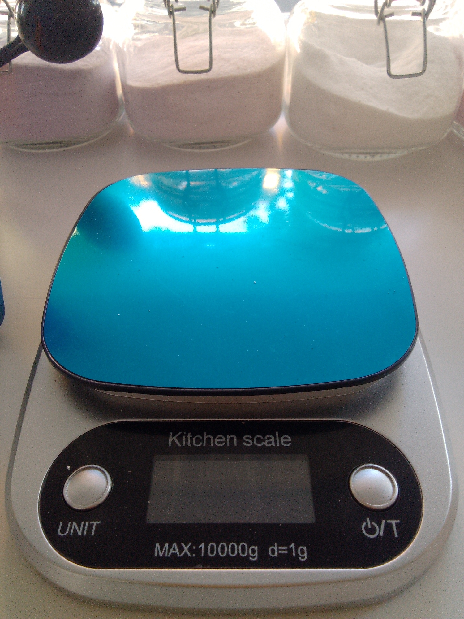 The image shows a close-up, high-angle view of a digital kitchen scale with a blue weighing platform. The scale is silver and black, with buttons labeled "UNIT" and a power/tare symbol. A digital display shows "MAX: 10000g d=1g". Behind the scale, out of focus, are three glass jars with metal clasps, each containing a different powder. The jar on the far left contains a pinkish powder, the middle jar contains a lighter pink powder, and the far-right jar contains white powder. A black scoop is partially visible in the far-left jar. The scene is brightly lit, likely indoors, on a flat, light-colored surface. The image does not provide any information about the city of Trenggalek, Indonesia, or any people.