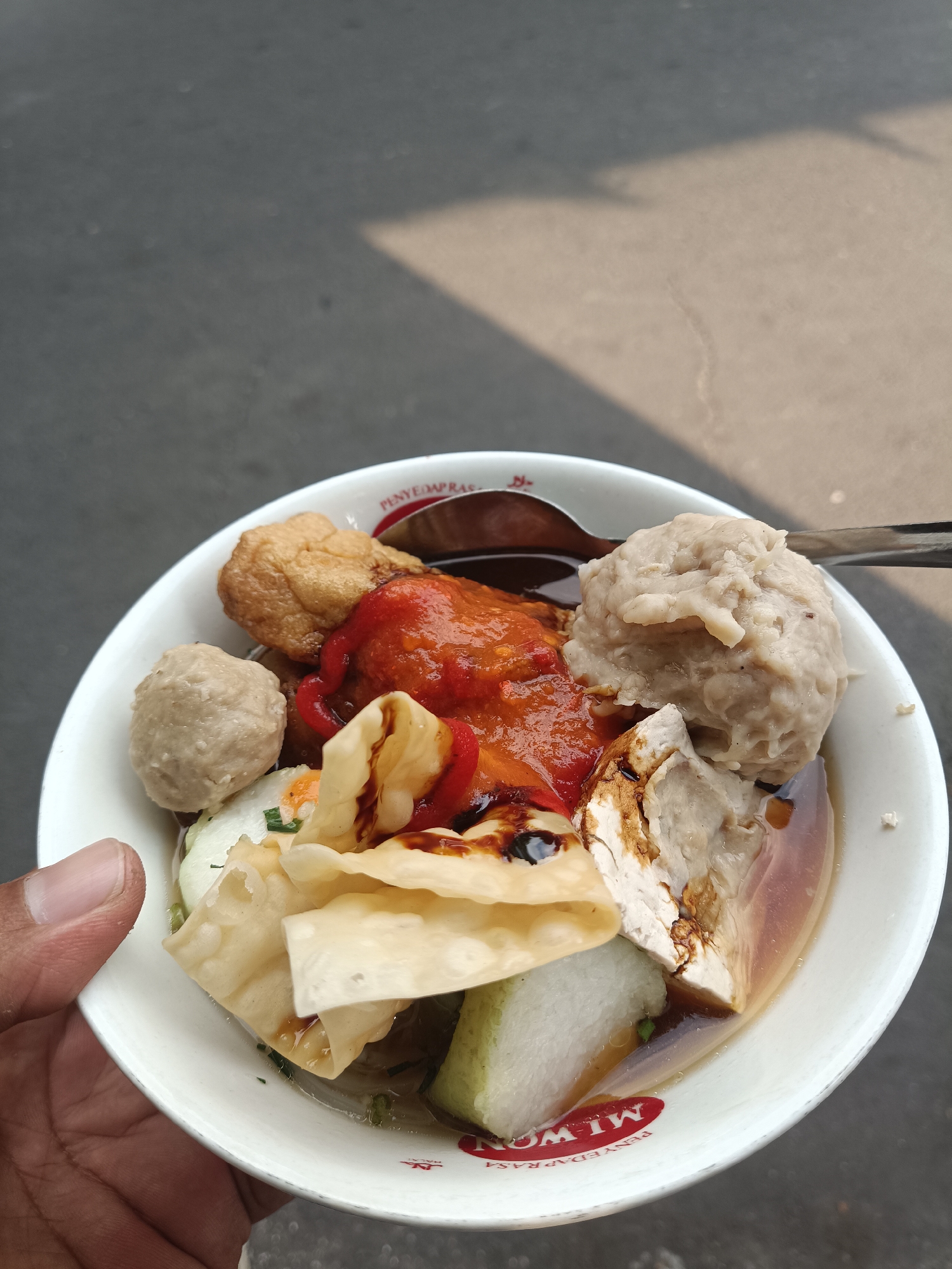 A close-up, overhead shot shows a white bowl of bakso, an Indonesian meatball soup, held by a person's hand. The bowl contains several large, light-colored meatballs, a piece of fried tofu, a fried wonton wrapper, and slices of cucumber, all submerged in a dark broth. A dollop of red chili sauce is visible on top, along with a drizzle of dark sauce. A spoon is resting in the bowl. The bowl has red writing around its base, which includes the word "MILWA" and "PENYE." The background is a blurred asphalt surface with a shadow from sunlight. The scene appears to be an outdoor setting in Jember, Indonesia, suggested by the branding on the bowl.