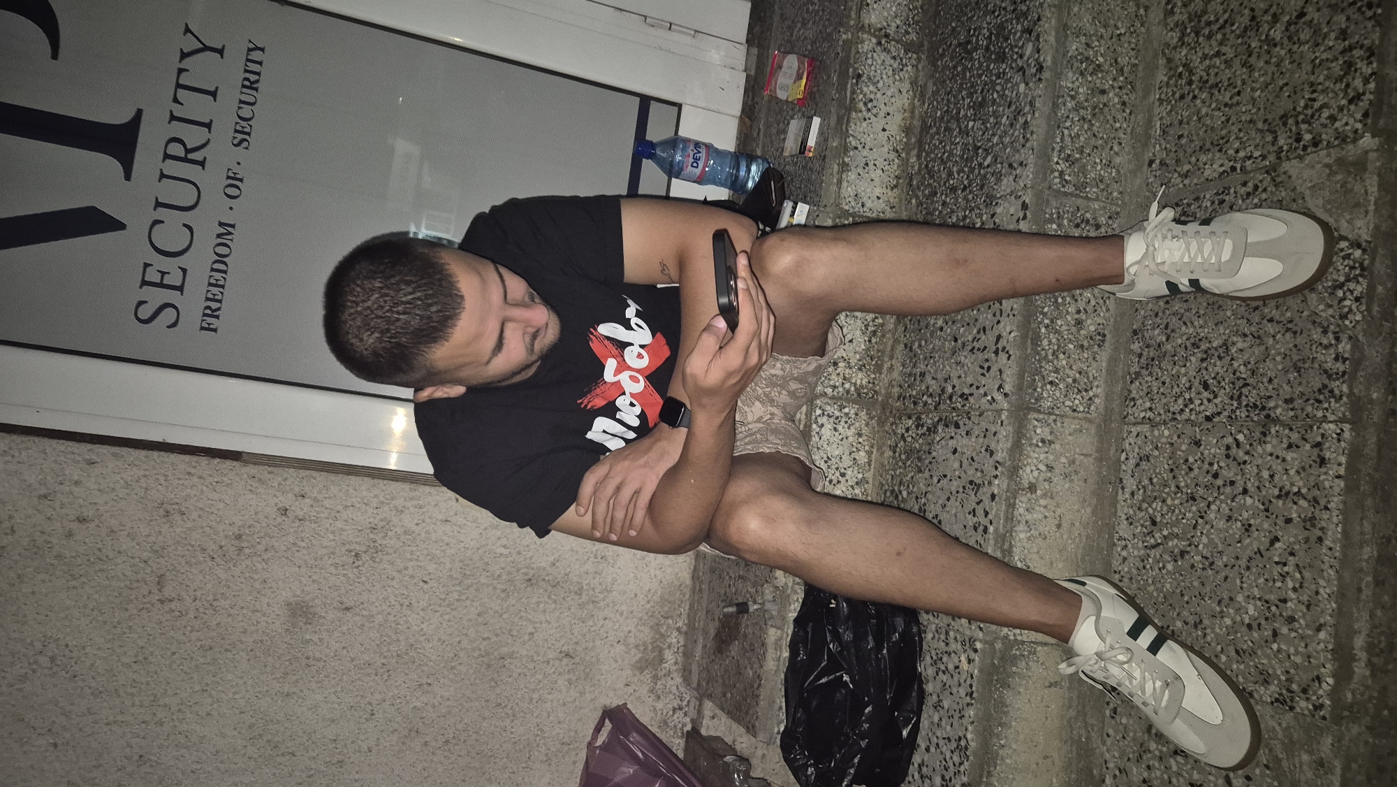 A young man sits on concrete steps outside a building with a sign that reads "SECURITY FREEDOM OF SECURITY". He is wearing a black t-shirt with a red "X" over the word "Любов" (Love), patterned shorts, and white sneakers. He is looking at his phone, which he holds in his hands. To his right, on the steps, are a bottle of water, a pack of cigarettes, and a lighter. To his left, a black trash bag and a purple plastic bag are visible. The background shows a rough concrete wall and the glass door of the building. The scene appears to be captured at night.
