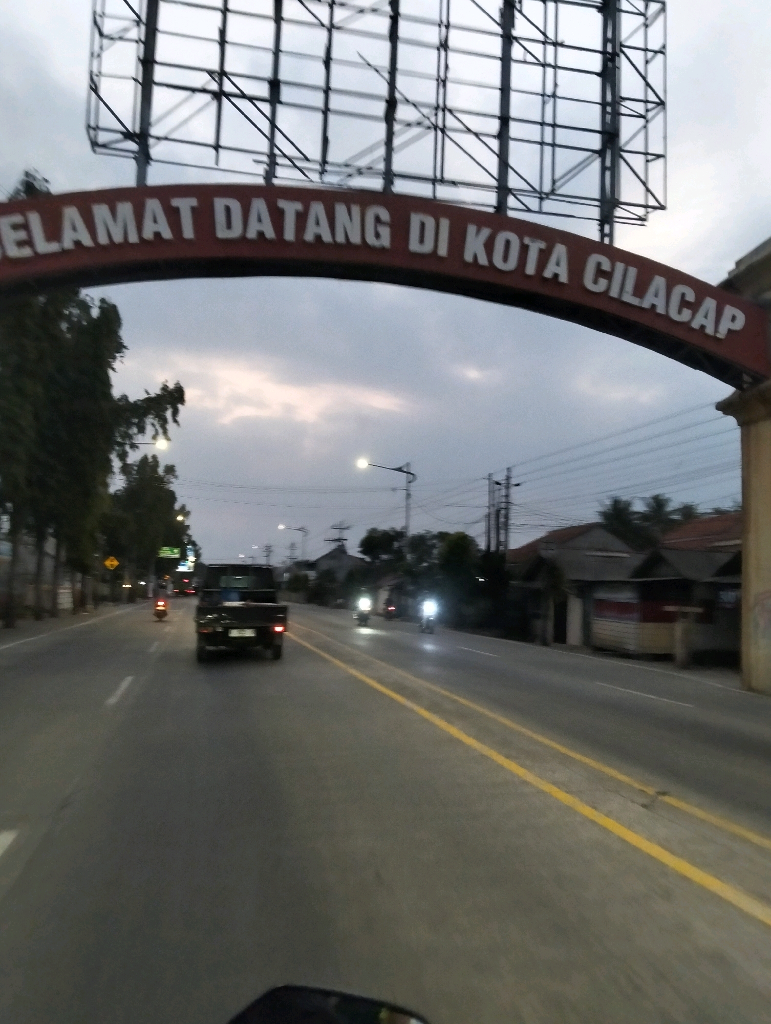 An archway with the text "SELAMAT DATANG DI KOTA CILACAP" (Welcome to the City of Cilacap) spans a road at dusk. A black pickup truck is driving away from the camera on the right side of the road. Several motorcycles are also present, with one clearly visible on the left side of the road, also moving away from the camera. The sky is overcast with a hint of light in the distance. Trees line the left side of the road, and buildings are visible on the right. The road has yellow lines marking the lanes. The overall scene suggests an entrance into the city of Cilacap, Indonesia, likely on a main thoroughfare.