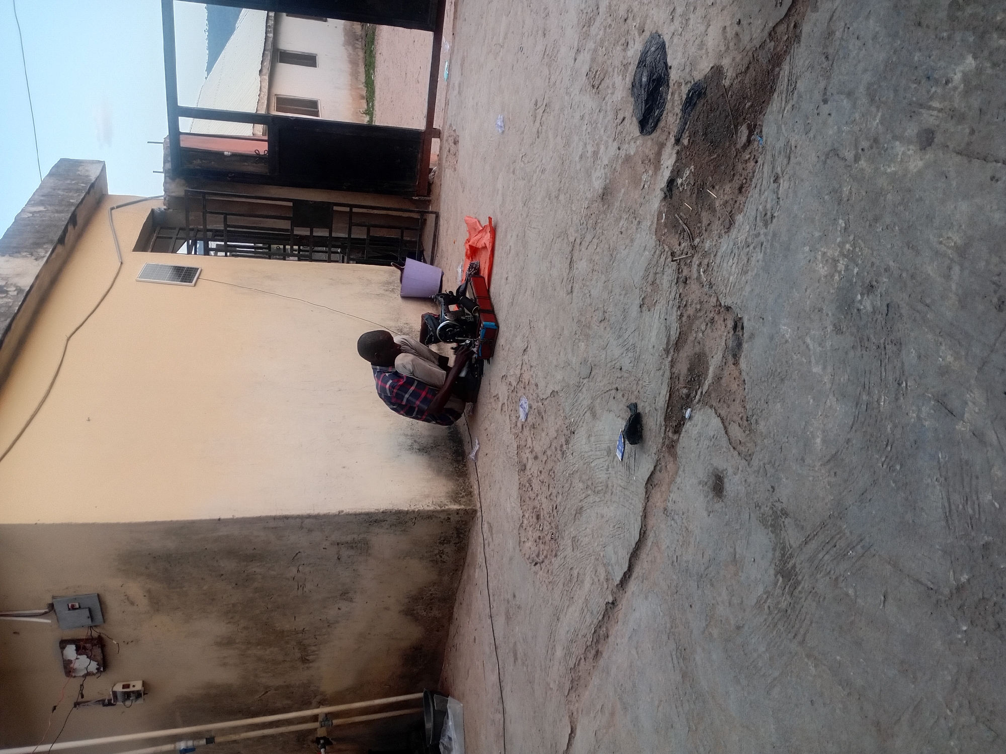 The image captures an outdoor scene in Lokoja, Nigeria, likely a courtyard or an informal workspace, under clear daytime conditions. A man is the central subject, seated on the ground and intently operating a traditional black sewing machine. He is wearing a plaid shirt and light-colored trousers. An orange mat or fabric is placed on the ground beneath the sewing machine, and a purple bucket rests nearby.
The surroundings feature light-colored building walls. On one wall, a small, rectangular solar panel is visibly mounted, suggesting a power source. Below it, several exposed electrical components, including small gray and brown boxes with wires, are affixed to the wall. In the background, metal bars cover windows, and another building with a corrugated metal roof is visible against a clear sky. The ground consists of a mixture of uneven concrete and dirt, with some scattered debris, including a black plastic bag and small pieces of paper. The overall scene depicts a small-scale, outdoor tailoring or repair operation, highlighting resourcefulness in a functional, basic environment. There is no visible text in the image.