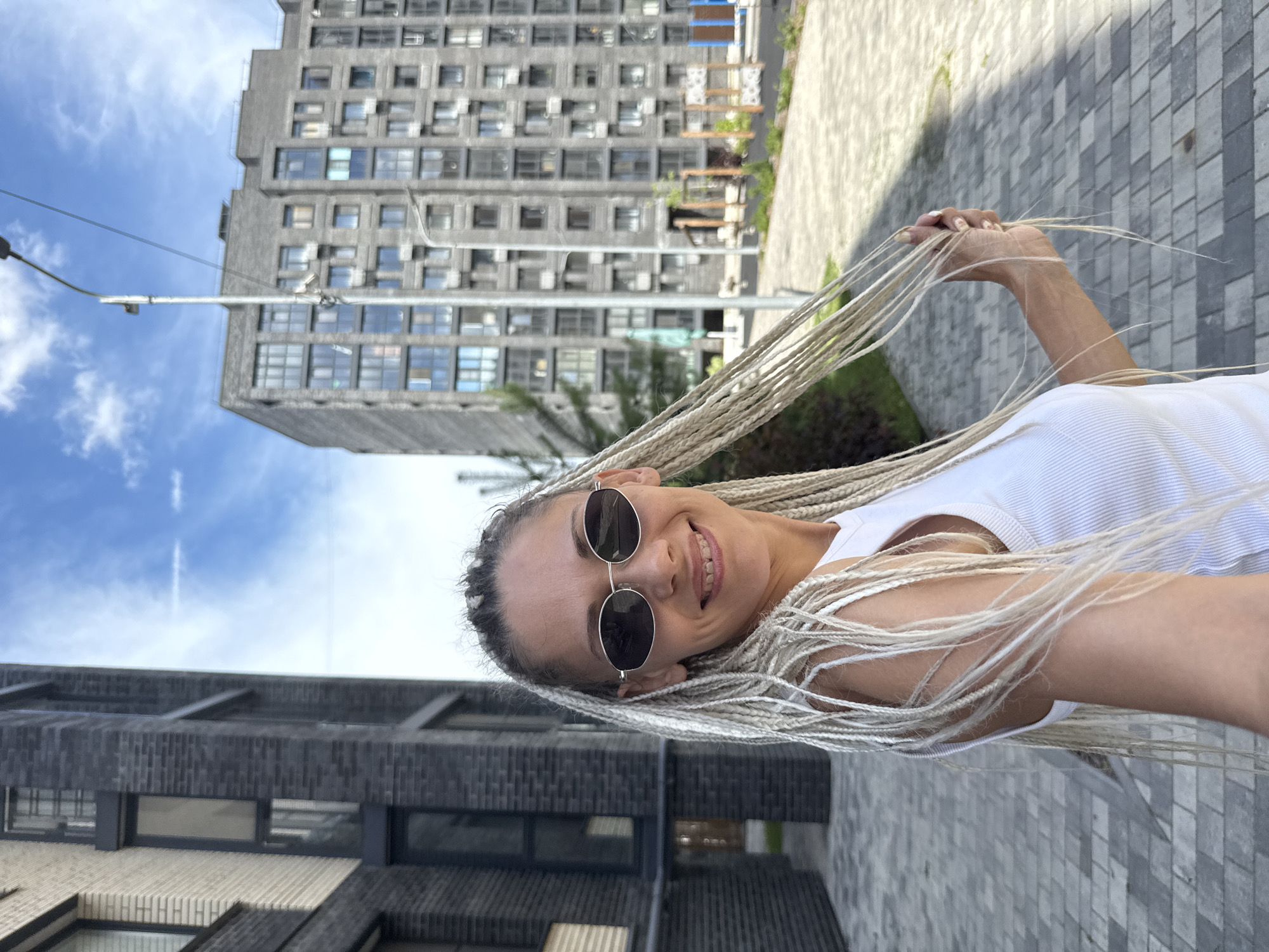 A medium shot captures a woman with blonde box braids and sunglasses, smiling at the camera. She is wearing a white tank top and stands on a paved walkway in front of modern, grey brick apartment buildings. A lamppost is visible in the background, and the sky is blue with some clouds. The location is identified as Rusanovo, Russia.