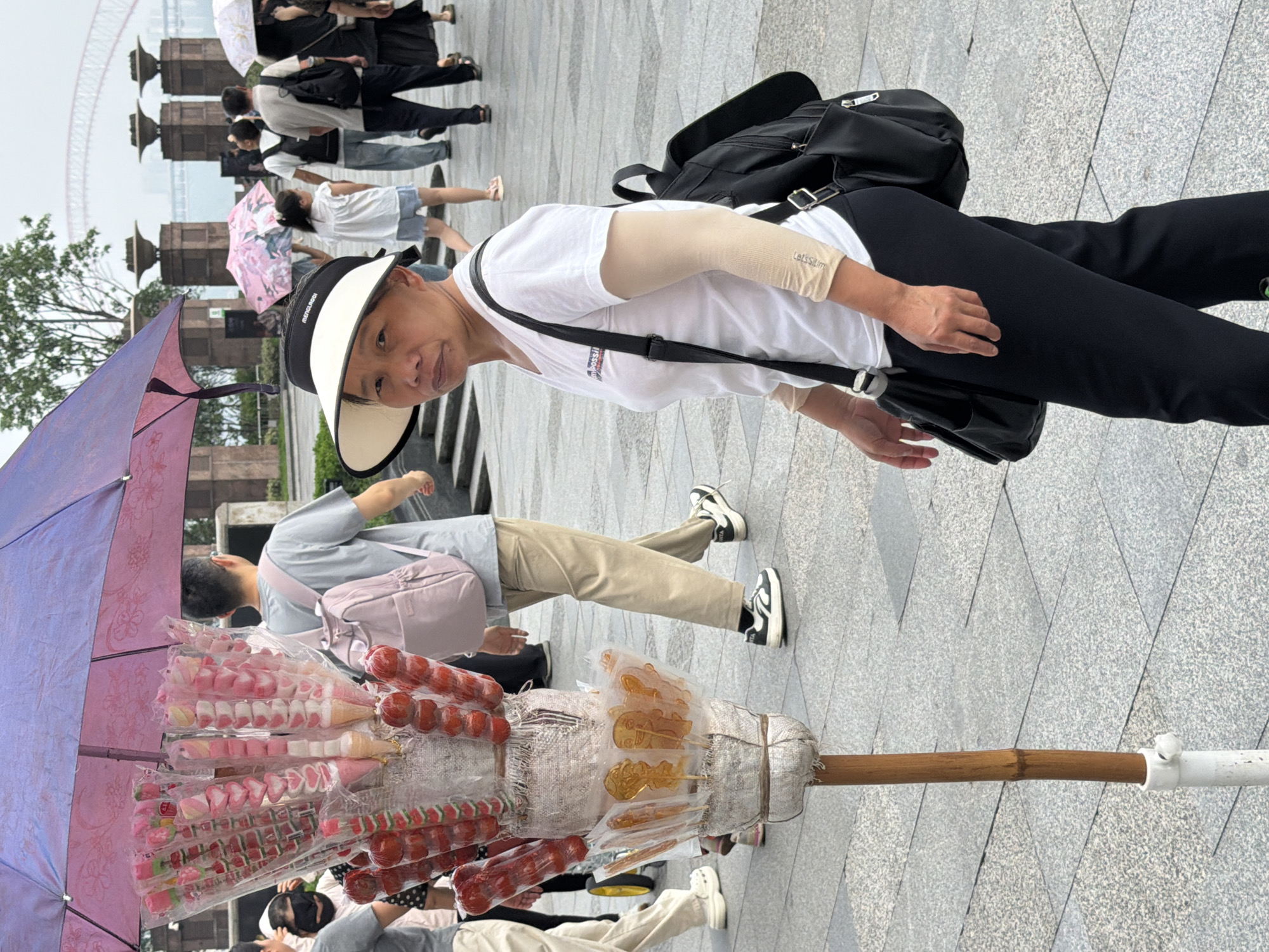 The image shows a street vendor in Yuzhong District, China, selling candied fruits and marshmallows from a stand. The vendor is a middle-aged woman wearing a white t-shirt, black pants, and a wide-brimmed hat. She has a black backpack and a black shoulder bag. In the background, several people are walking, some holding umbrellas, suggesting it might be raining or the sun is strong. The street is paved with gray tiles, and there are some structures and trees in the background. The candy stand is decorated with a purple umbrella and displays various colorful sweets on sticks.