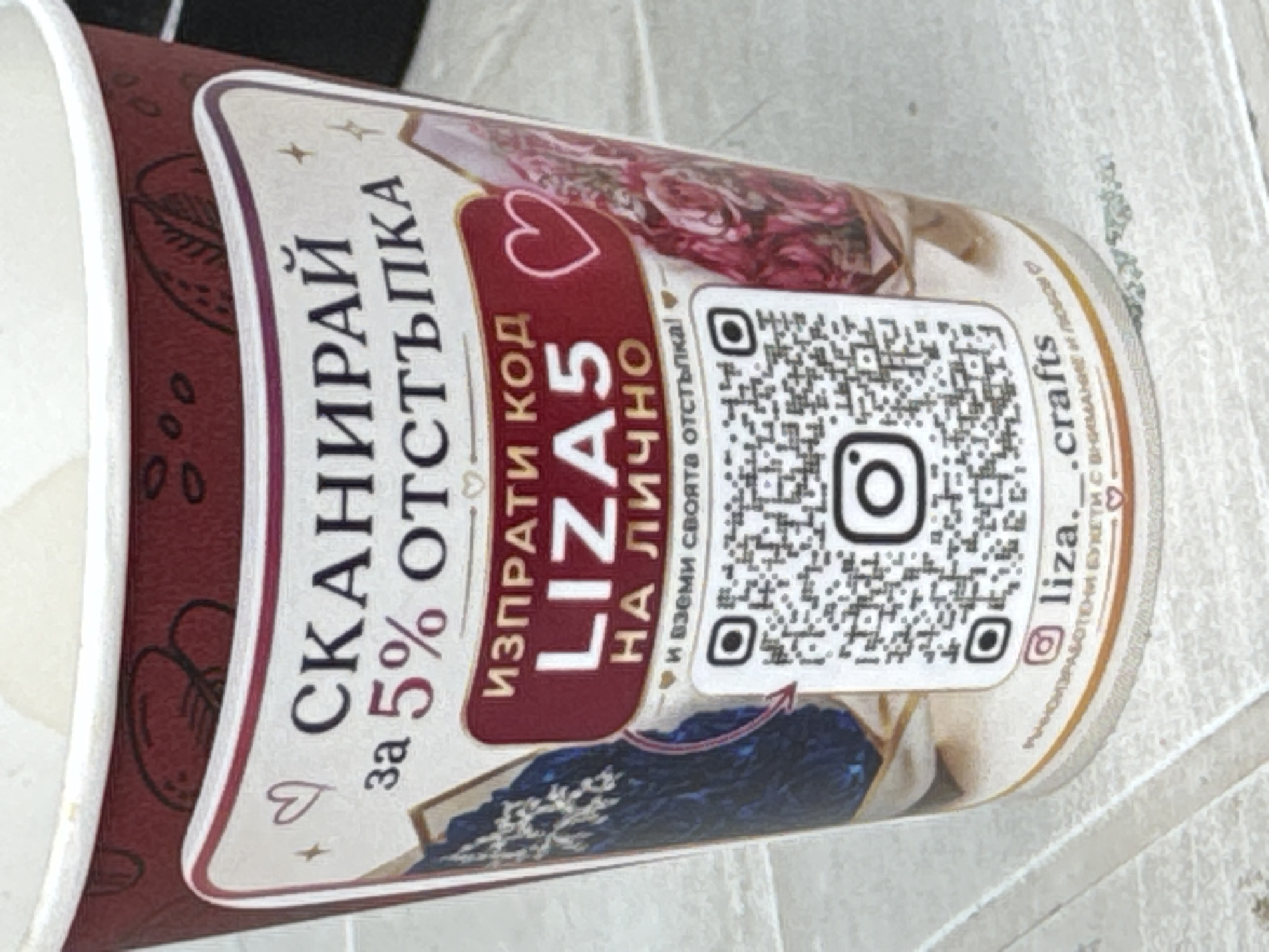 The image shows a close-up of a paper cup, likely for a beverage, with various graphics and text. The cup is dark red with white and gold accents. Prominently displayed is a QR code and an Instagram logo. Above the QR code, there is text in what appears to be Cyrillic script, including the word "СКАНИРАЙ" (SCAN) and "5% ОТСТЪПКА" (5% DISCOUNT). Below this, the code "LIZA5" is highlighted in large white letters on a dark red background. Further text below indicates "НА ЛИЧНО" (FOR PERSONAL) and "и вземи своята отстъпка!" (and get your discount!). At the bottom of the cup, an Instagram handle ".liza._crafts" is visible, followed by more Cyrillic text that is partially obscured. The cup is placed on a light-colored surface, possibly a table or countertop, with visible texture. The background is blurred, but suggests an indoor environment. There are no people visible in the image. The specific location in the city of Sredets, Bulgaria, cannot be determined from the visual information provided.