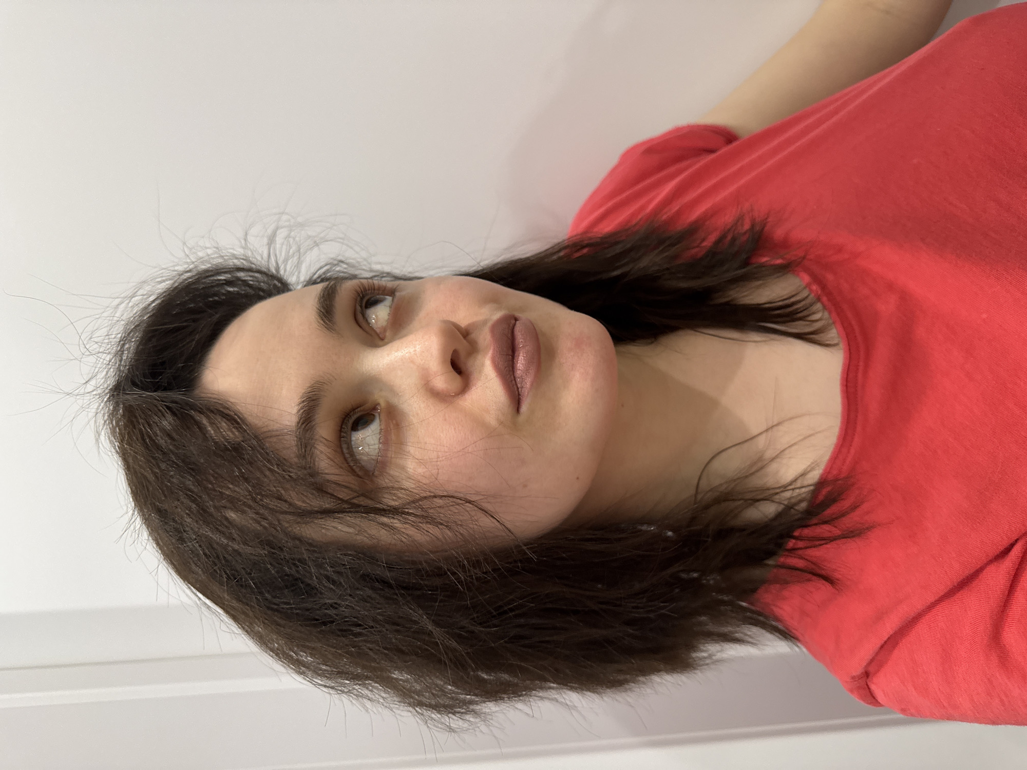 The image is a close-up portrait of a young woman looking upwards and to her right. She has dark, medium-length hair that appears slightly windswept, framing her face. She wears a plain red t-shirt. Her complexion is fair, and her lips are a muted pink or mauve color. Her eyes are light brown, with a noticeable sheen, and her eyebrows are dark and full. The background is a simple, light-colored (likely white or off-white) wall, with faint horizontal lines indicating a paneled or textured surface, suggesting an indoor setting in Izhevsk, Russia. The lighting is soft and diffused, casting gentle shadows. Her expression appears calm and contemplative. No text is visible in the image.