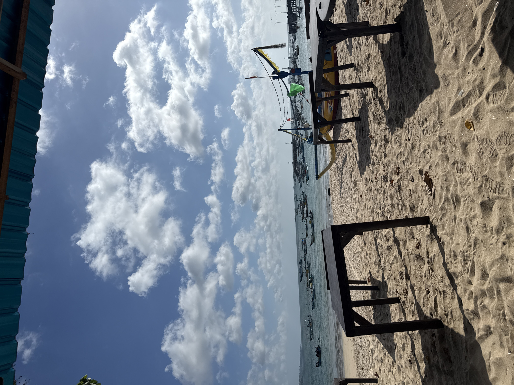 The image shows a beach scene, likely in Kuta, Indonesia, given the presence of traditional outrigger boats on the water. The sky is blue with scattered clouds. Tables are placed on the sand, casting shadows. In the background, numerous boats are anchored in the sea, some with decorative flags. The scene is predominantly outdoors with natural light. The presence of beach furniture suggests a tourist or recreational area. The overall environment is coastal.