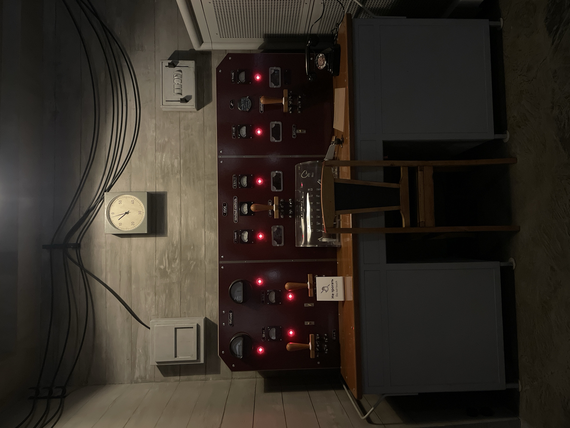 The image shows an interior space with a vintage, possibly Cold War-era control panel. The wall is textured, likely concrete or painted to resemble it. Various electrical conduits and wires run along the wall, suggesting a functional or industrial setting. A clock is mounted on the wall, displaying a time of around 11:50. Several control panels are mounted on the wall, featuring gauges, switches, and indicator lights. A sign with Russian and English text saying "Do not touch" is attached near one of the panels. A telephone is visible on the top panel. To the right of the control panel is a desk-like structure with drawers or cabinets.
Given the style of the control panel, the general aesthetic, and the presence of Russian text, it is plausible that the image was taken at Bunker-42 on Taganka Square in Moscow, Russia. Bunker-42 is a former secret Cold War command center that has been converted into a museum.