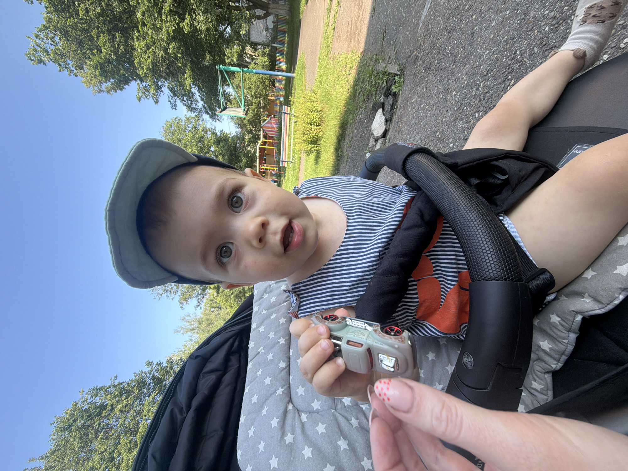 In Chaikovsky, Russia, on a sunny day, a baby is sitting in a stroller, holding a toy car. The baby is wearing a striped shirt and a cap. A person's hand is visible in the foreground, near the baby's toy. The stroller is parked on a paved surface, with grass and trees in the background. A playground structure is visible further in the background.