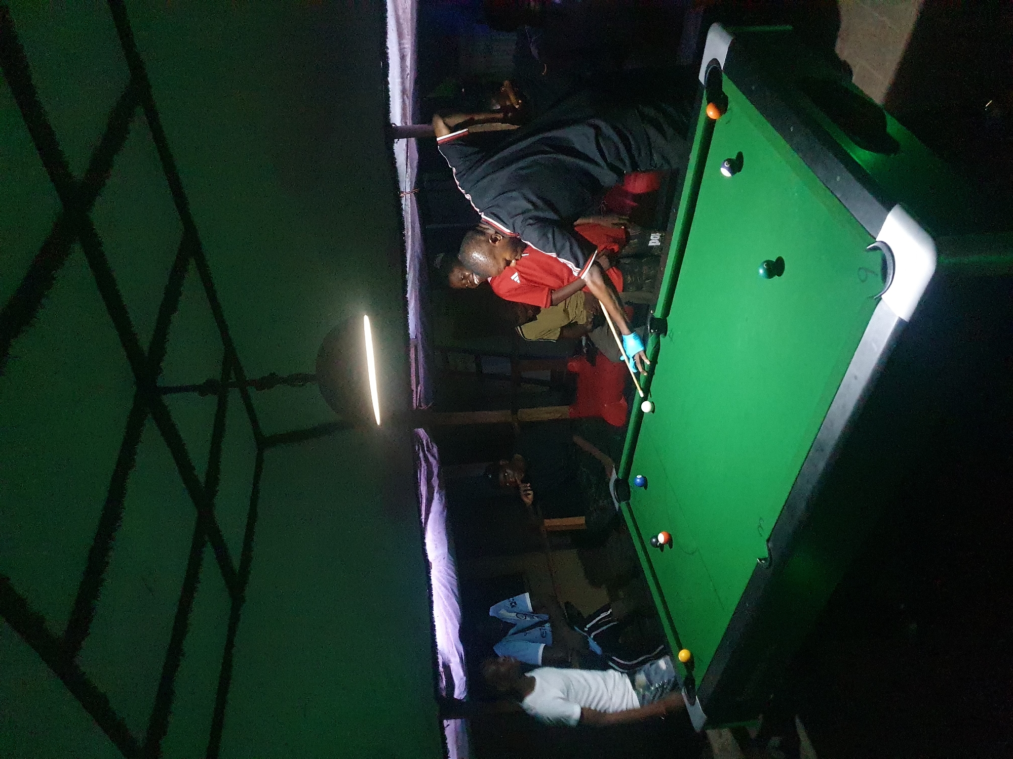 The scene depicts an indoor establishment, possibly a bar or recreational space, in Lokoja, Nigeria, given the available location information. Two individuals are positioned at a green pool table with multiple pool balls arranged on it. One person is actively playing pool, with a pool cue in hand and wearing what appears to be a blue glove. They are clad in a red shirt with white accents. Another individual, dressed in black, is observed close to the pool player, potentially observing or participating in the game. A third person is lying on a nearby couch, wearing a white shirt. Overhead, a light fixture illuminates the pool table and surrounding area. The walls of the space are visible, and there is a structural design element, potentially a roof support or decorative feature, visible in the upper left corner. The overall lighting appears to be dim, contributing to a relaxed or intimate atmosphere.