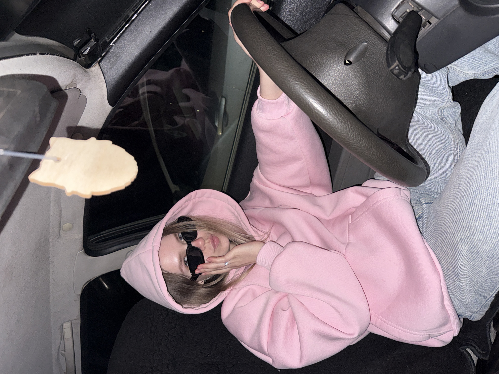 A young woman is depicted inside a car, lying on her side, possibly across the front seats. She is wearing a light pink hooded sweatshirt with the hood pulled up, and black sunglasses obscure her eyes. Her light blonde hair is visible around her face. One hand, adorned with a ring, rests near her chin as she poses, looking directly at the camera with slightly parted lips. Her other hand is holding onto the steering wheel. She is also wearing light blue jeans.
To the left of the frame, a light-colored, irregularly shaped object resembling a cookie or biscuit is held on a thin metal fork or skewer, extending into the shot. The interior of the car is visible, including parts of the dashboard, the dark steering wheel, a door panel, and the dark fabric of a car seat. The lighting in the scene is dim, suggesting an indoor setting or nighttime. There is no visible text within the image. The location context provided for this image is Zhigalki, Russia.