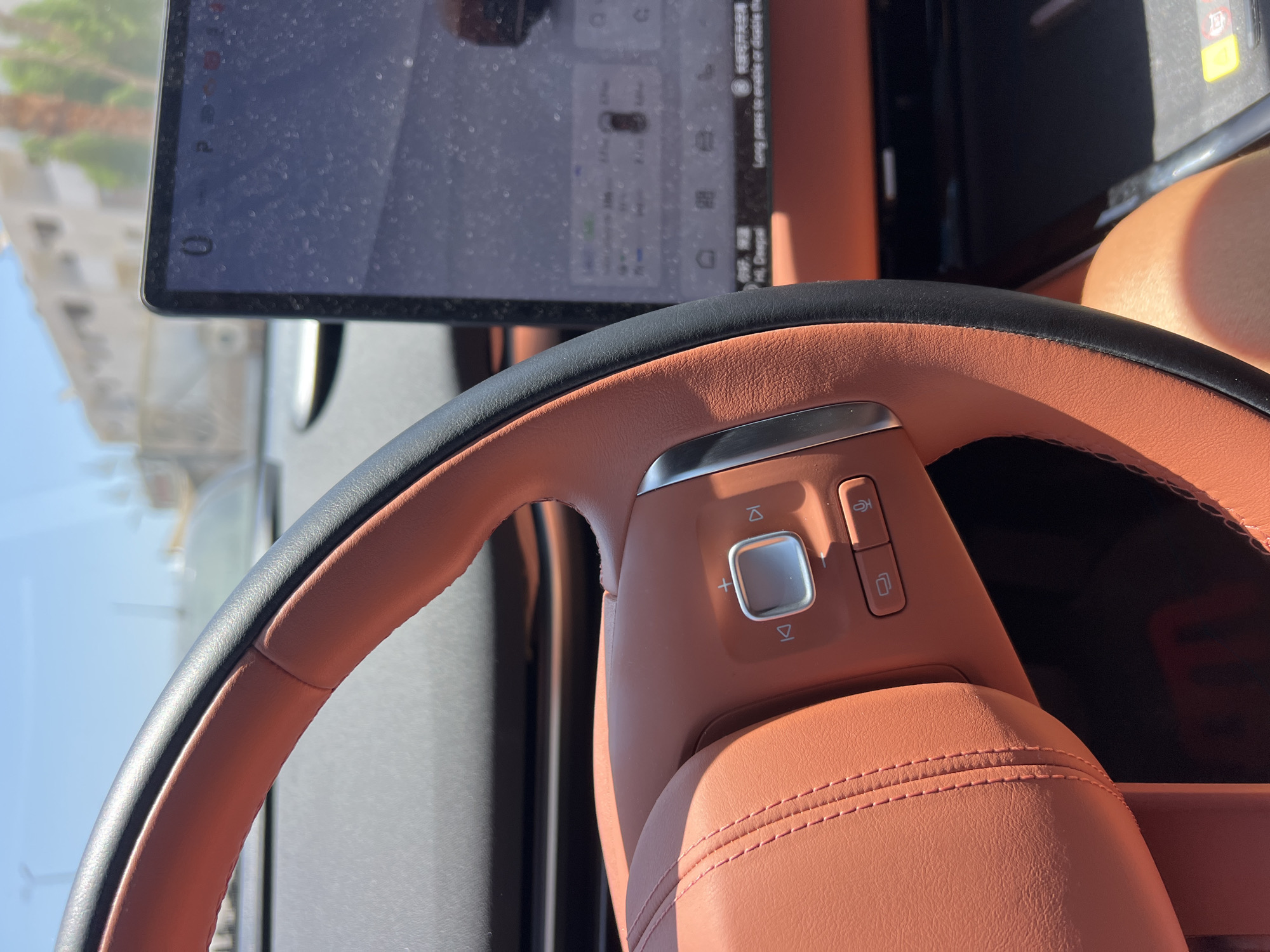 The image shows the interior of a car, specifically the steering wheel and a touchscreen display. The steering wheel is covered in orange leather with black stitching and a black rim. On the steering wheel, there are several buttons, including a circular button with a plus sign, a play/pause button, and navigation buttons. The touchscreen display, located to the right of the steering wheel, shows a car icon and some menu options, but the details are difficult to discern due to dust on the screen. The background visible through the car's windows suggests an outdoor environment with buildings and trees, indicating a sunny day. The lower part of the image reveals a person's leg. Based on the presence of distinct architecture in the background and the overall atmosphere, the location is identified as Sharm El Sheikh, Egypt.