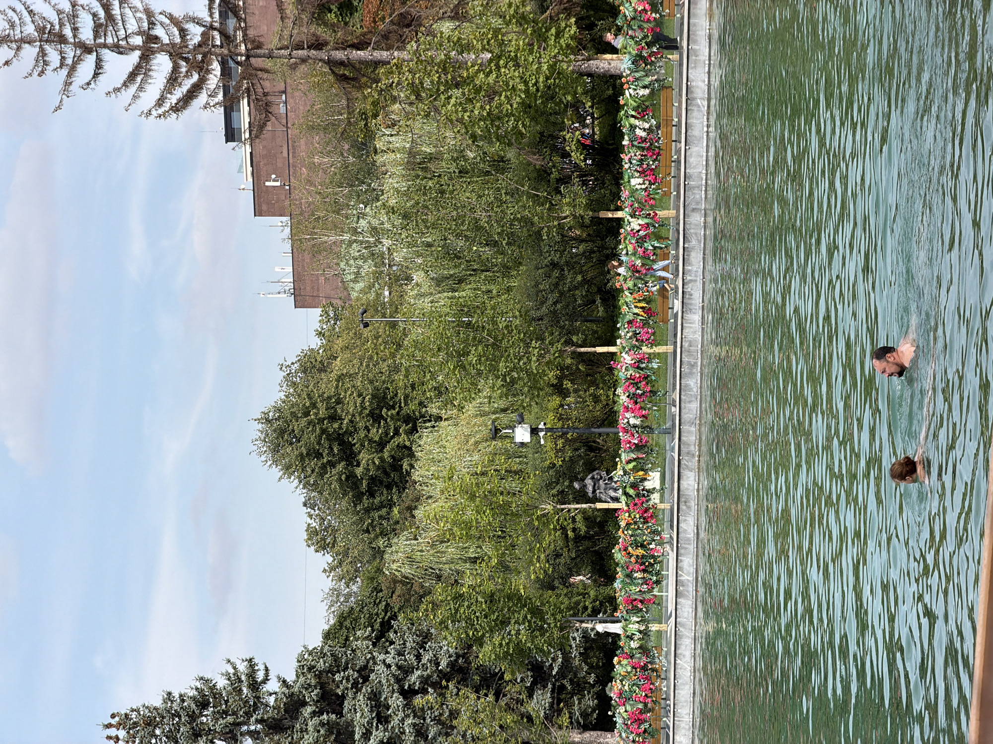 The image depicts a scene at the Patriarch's Ponds in Moscow, Russia. In the foreground, two individuals are swimming in a body of water. Behind them, a decorative fence adorned with numerous colorful flowers separates the water from a park area. This park features several trees, including a tall, dry pine tree on the right, and a dense array of green foliage. In the background, a red brick building with a modern top section is visible, along with a clear blue sky with scattered clouds. Several people are seen walking or standing in the park area.