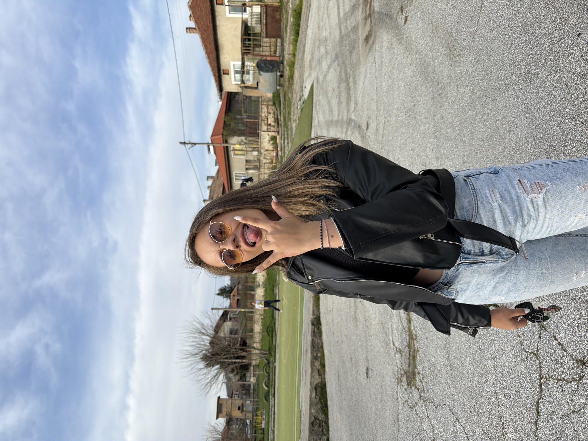 A young woman with long, light brown hair stands outdoors on a paved surface. She wears round, gold-rimmed sunglasses, a black leather jacket, and ripped light blue jeans. She extends her left hand, making a gesture with her fingers while sticking out her tongue. In her right hand, she holds keys. The background features buildings with orange-tiled roofs, a green field with yellow lines, and a blue sky with scattered clouds. The image was likely taken near the football field and community center in Popintsi, Bulgaria.