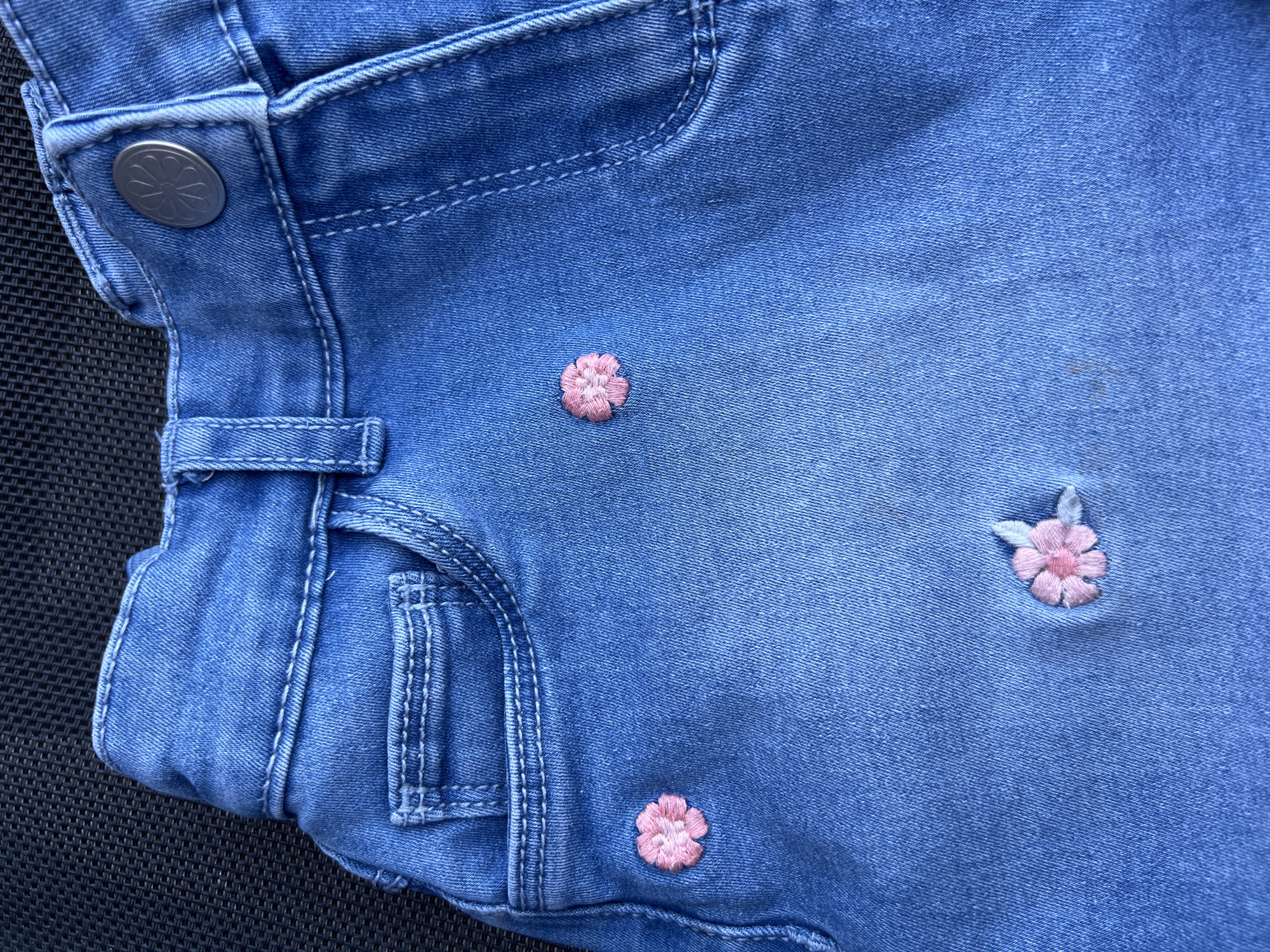 The image displays a close-up view of a pair of light blue denim shorts, likely for a child, with embroidered pink flowers. The shorts are textured with the characteristic weave of denim and feature visible stitching, a pocket, and a button at the waistband. The flowers are small and delicate, with a few scattered across the fabric. The background is a dark, textured surface, possibly a rug or furniture. The scene is indoors and does not provide any information about the location in Eretria Municipal Unit, Greece.