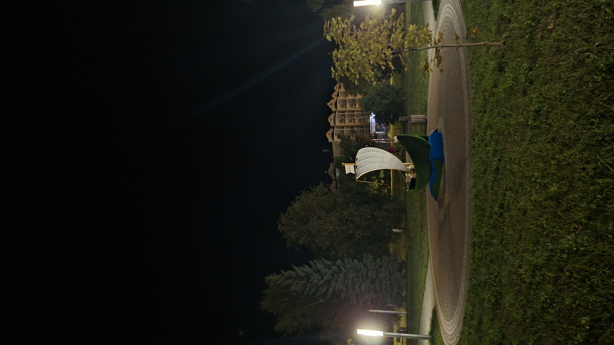 In Myadzyel, Belarus, at night, a playground feature resembling a green sailboat sits on a blue base in a park. The sailboat has a white, perforated sail and is positioned on a circular paved area surrounded by grass. Tall evergreen trees and other deciduous trees form a backdrop, with a multi-story building visible in the distance. Two modern lampposts illuminate the park. The sky is dark and clear, with no visible celestial bodies.