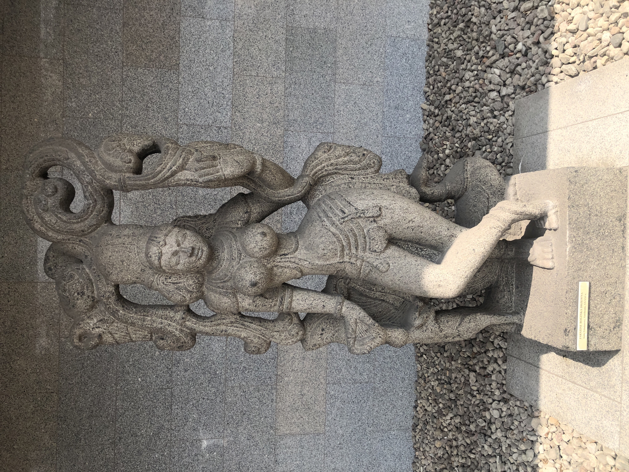 A stone sculpture of a female figure is displayed outdoors against a textured grey wall. The sculpture, likely an ancient artifact, depicts a woman in a dynamic pose, with detailed clothing and jewelry. The figure has distinct facial features and elaborate headdresses. The sculpture rests on a stone pedestal. The ground in front of the pedestal is covered with gravel and then a paved area. A small plaque is visible at the base of the pedestal with text in a foreign language. This sculpture is located in the Warsaw Zoological Garden in Warsaw, Poland, specifically within the Asian Rainforest exhibit, which aims to recreate the atmosphere of a tropical jungle with lush vegetation and ancient artifacts.