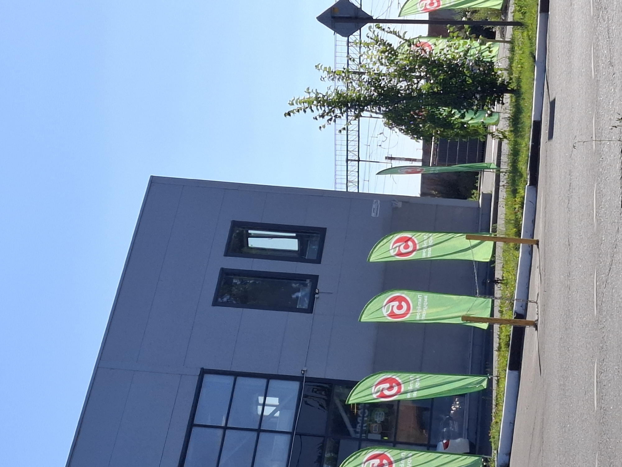 The image shows the exterior of a modern building with a light gray facade and large windows. In front of the building, several tall, green flags with a red circular logo are displayed on poles. A small, leafy tree stands to the right of the flags, partially obscuring a metal fence and some power lines in the background. The scene appears to be a sunny day, with a clear blue sky. The area in front of the building is paved, suggesting a commercial or public space.