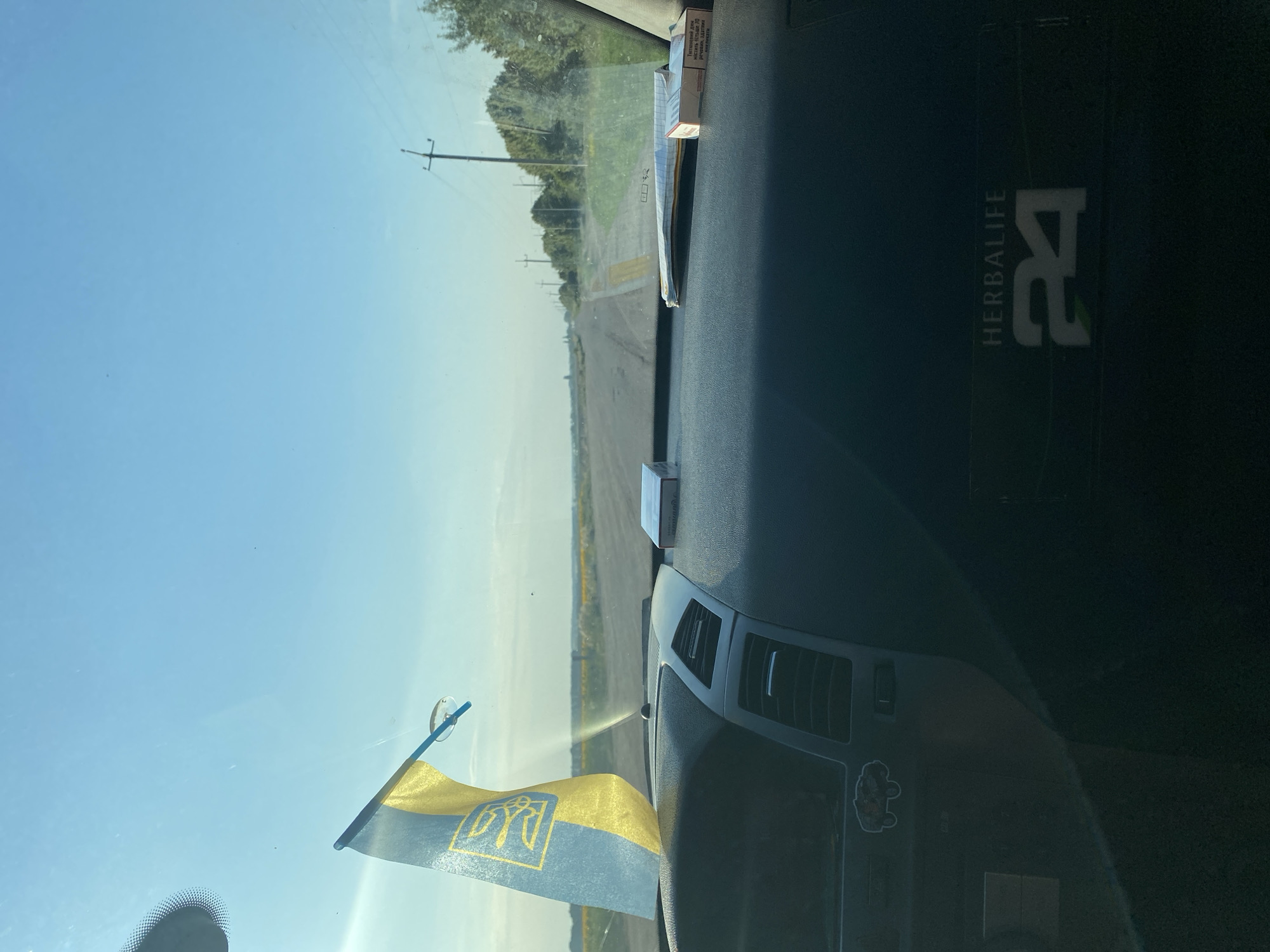 A first-person perspective shot from inside a car, looking through the windshield at a rural road. The dashboard is visible in the foreground, with a Ukrainian flag attached to the windshield on the left. Two packs of cigarettes and a folded piece of paper are on the dashboard. The road stretches into the distance, lined with trees and power lines. The sky is clear and blue. A Herbalife 24 logo is visible on the lower right of the dashboard. The scene is set in the city of Maiske, Ukraine, on a sunny day.