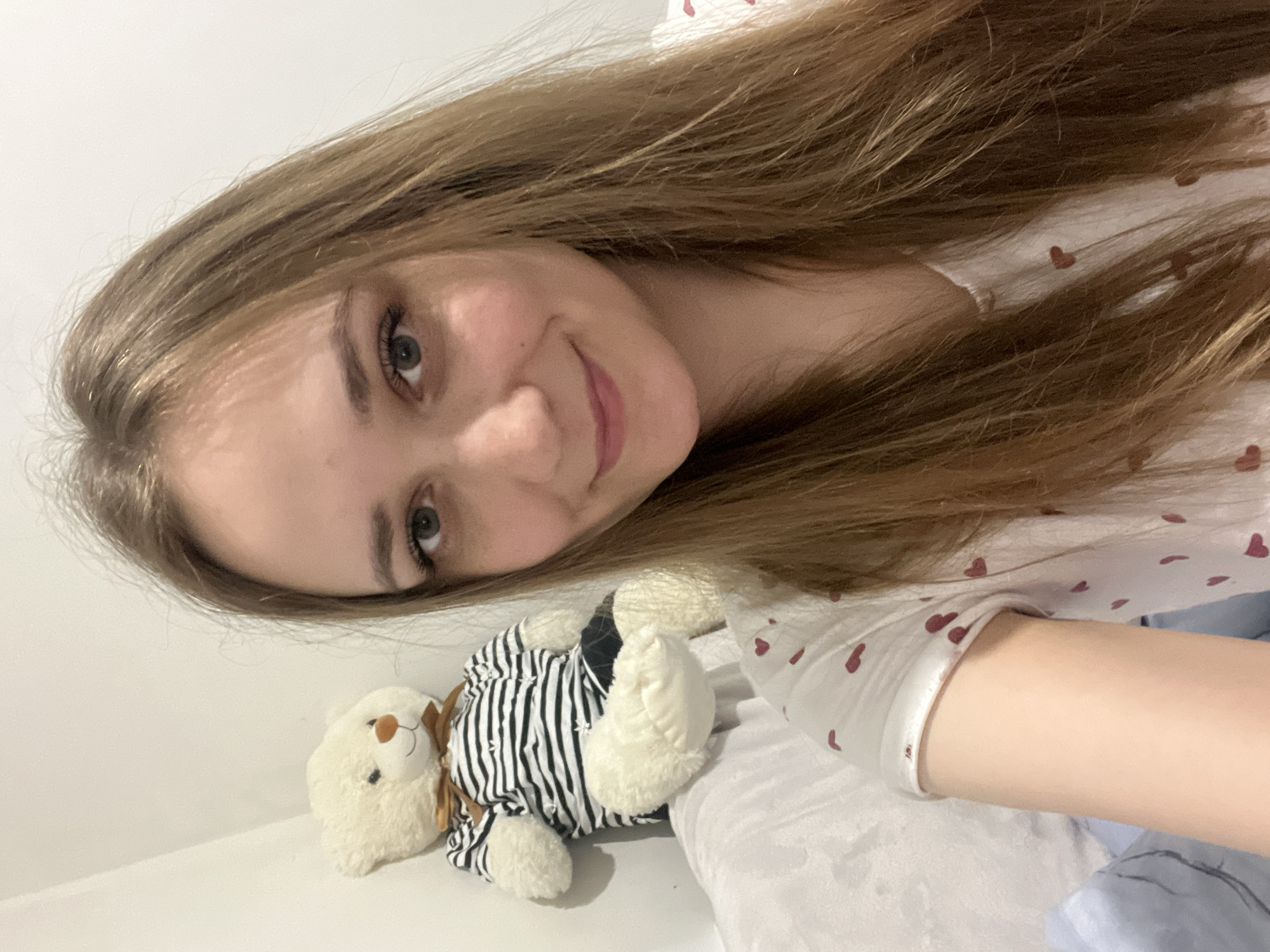 A young woman with long brown hair is lying on a white bed, smiling at the camera. Next to her is a white teddy bear wearing a black and white striped outfit and a brown bow tie. The woman is wearing a white shirt with small red hearts printed on it. The scene is indoors and well-lit. There is no specific information to determine the location of this photo, so I can't determine it is Verkhnyaya Pyshma, Russia.