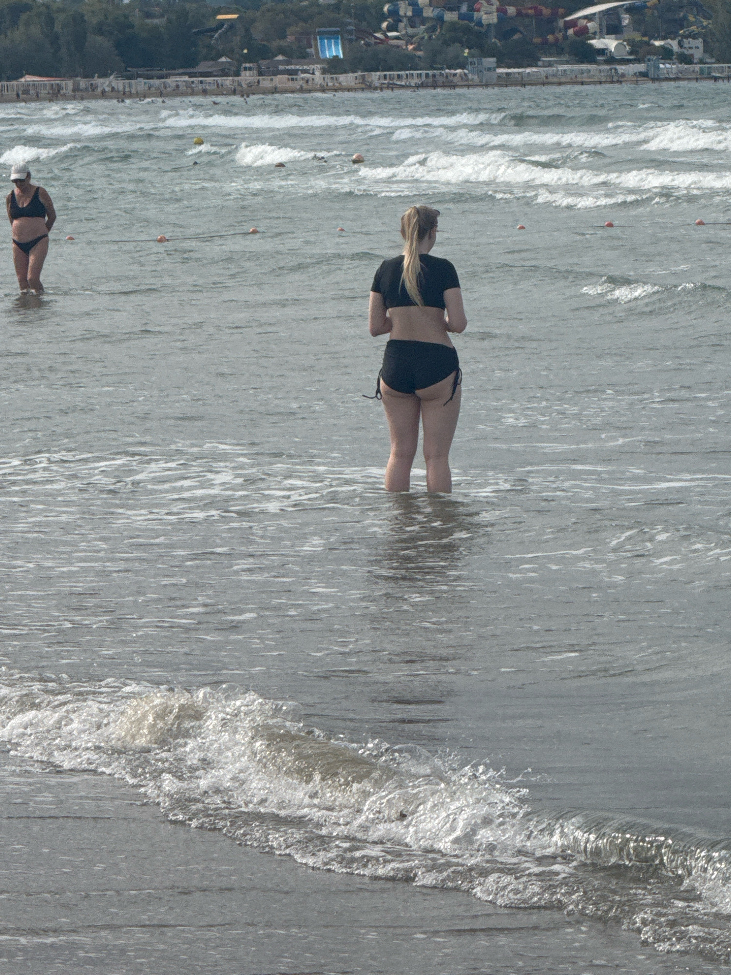 Two women are standing in shallow ocean water near a beach. The woman on the left is wearing a white cap and a black bikini. The woman on the right is wearing a black bikini top and bottoms. In the background, there are waves, buoys, and a distant shoreline with buildings and trees. The scene appears to be a public beach in Anapa, Russia, a resort city on the Black Sea.