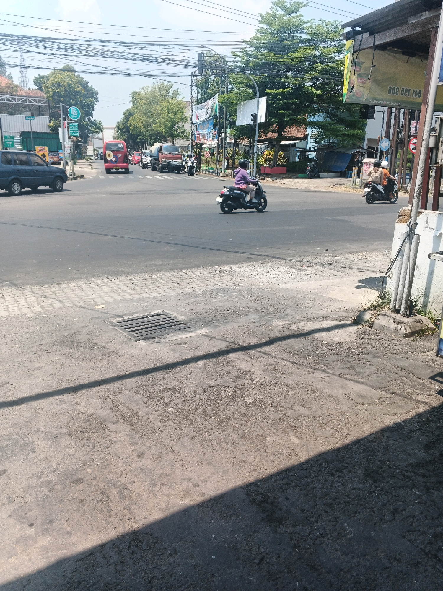 This is a street-level, eye-level shot of a busy intersection in Ungaran, Indonesia. The day is bright and sunny, with shadows cast across the asphalt. The foreground shows a concrete sidewalk with a metal grate drain. The mid-ground depicts the intersection itself, with traffic moving in various directions. A dark blue SUV is parked on the left. A red van is moving forward, followed by other vehicles. Two motorcycles are also visible; one is ridden by a person in a purple shirt and the other carries two people. In the background, trees, buildings, and utility poles with a dense network of wires are visible. A billboard with text is also present on the right side of the image. The overall scene suggests a typical urban environment with active traffic.