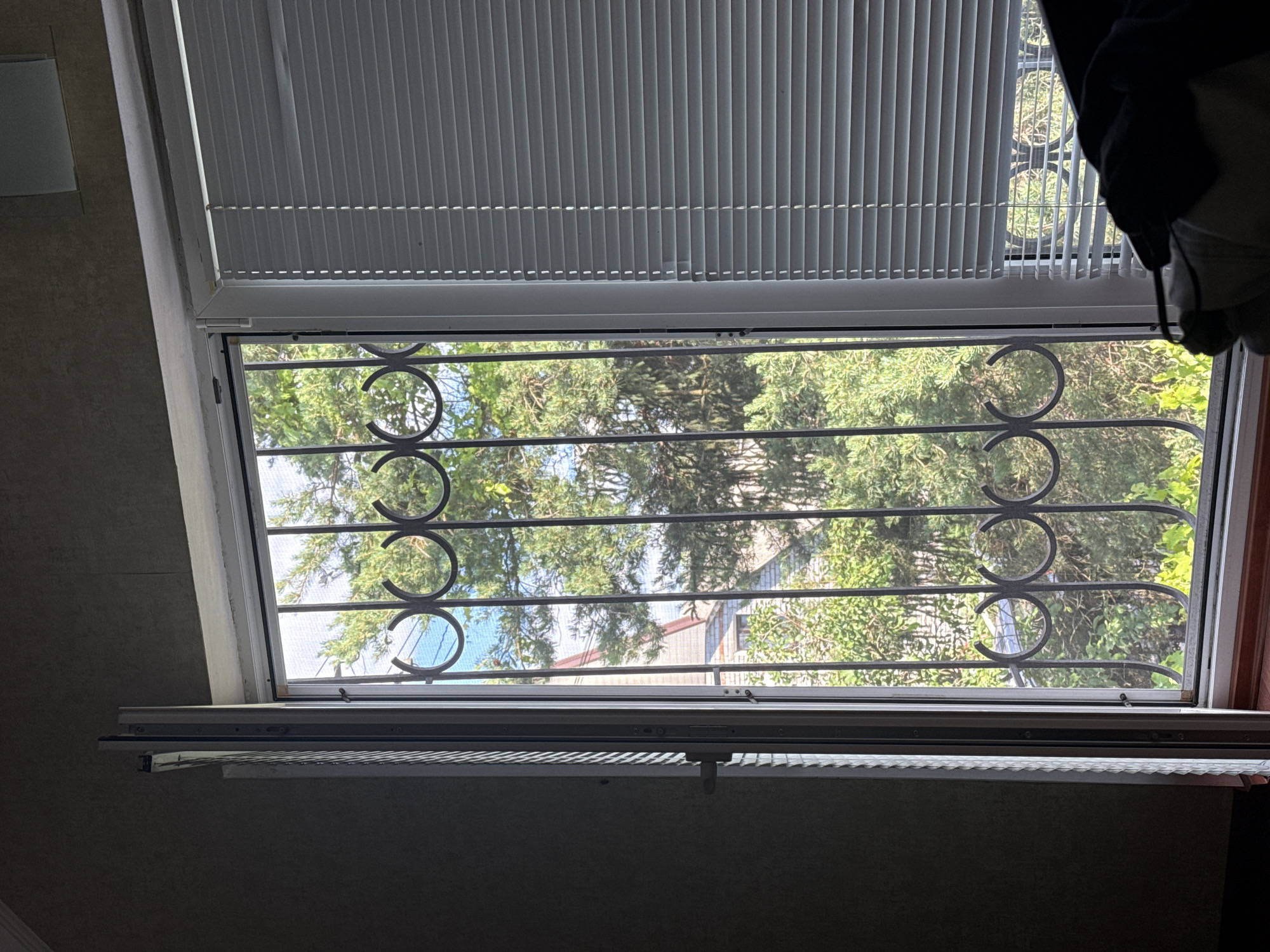 The image shows a view through a window in Obukhivka, Ukraine. The window is fitted with a metal security grate, which has a decorative pattern of concentric circles. Outside the window, there are trees with green foliage, and a building with a reddish-brown roof can be seen through the branches. The sky is visible in patches between the leaves, suggesting it is daytime. The top of the window is covered by white blinds, which are partially open, allowing light to enter the room. The interior of the room is dimly lit, with the wall on the left appearing to be textured and painted a neutral color. A dark curtain or piece of clothing hangs from the upper right corner of the frame, obscuring part of the window. The overall impression is of a residential setting, possibly a ground-floor window given the presence of the security grate. The time of day appears to be morning or afternoon, with the weather likely clear enough to allow for natural light to filter through the trees. No text is visible in the image.