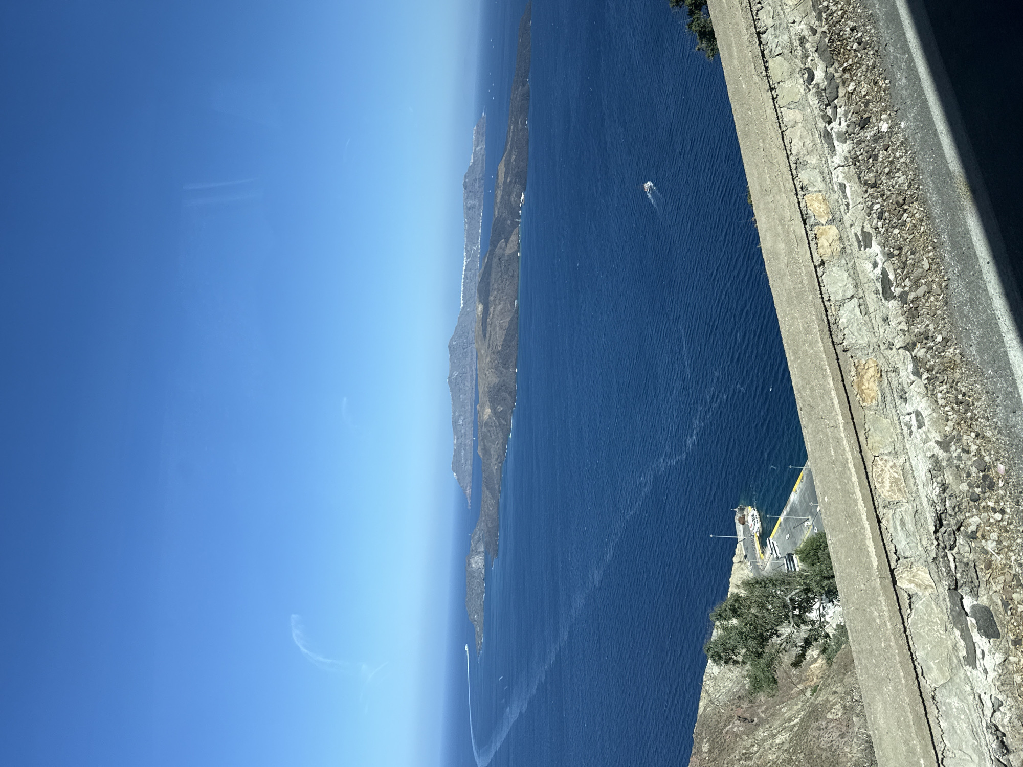 A high-angle shot captures a vast expanse of deep blue Aegean Sea under a clear, bright sky. In the foreground, a rough, stone-and-concrete retaining wall runs horizontally across the bottom of the frame, with a paved road visible below it. The wall's upper edge is lined with weathered concrete, and beneath it, a jumble of rocks and gravel is visible.
Beyond the wall, the sea dominates the scene. Several islands, appearing rugged and mountainous, are scattered across the water, their dark, rocky surfaces contrasting with the bright blue sea and sky. Some of these islands are lined with white buildings, suggesting a settlement. White wakes trail behind a small boat moving across the sea. In the mid-ground, a quay or harbor is visible, with a white ferry or tour boat docked alongside. A few other smaller boats are also present. Trees and sparse vegetation grow on the steep, rocky slopes leading down to the water.
The scene is likely from the caldera of Santorini, a volcanic island in Greece, possibly from a vantage point in Thira Municipal Unit. The distinctive geological formations and the presence of the caldera suggest this location.