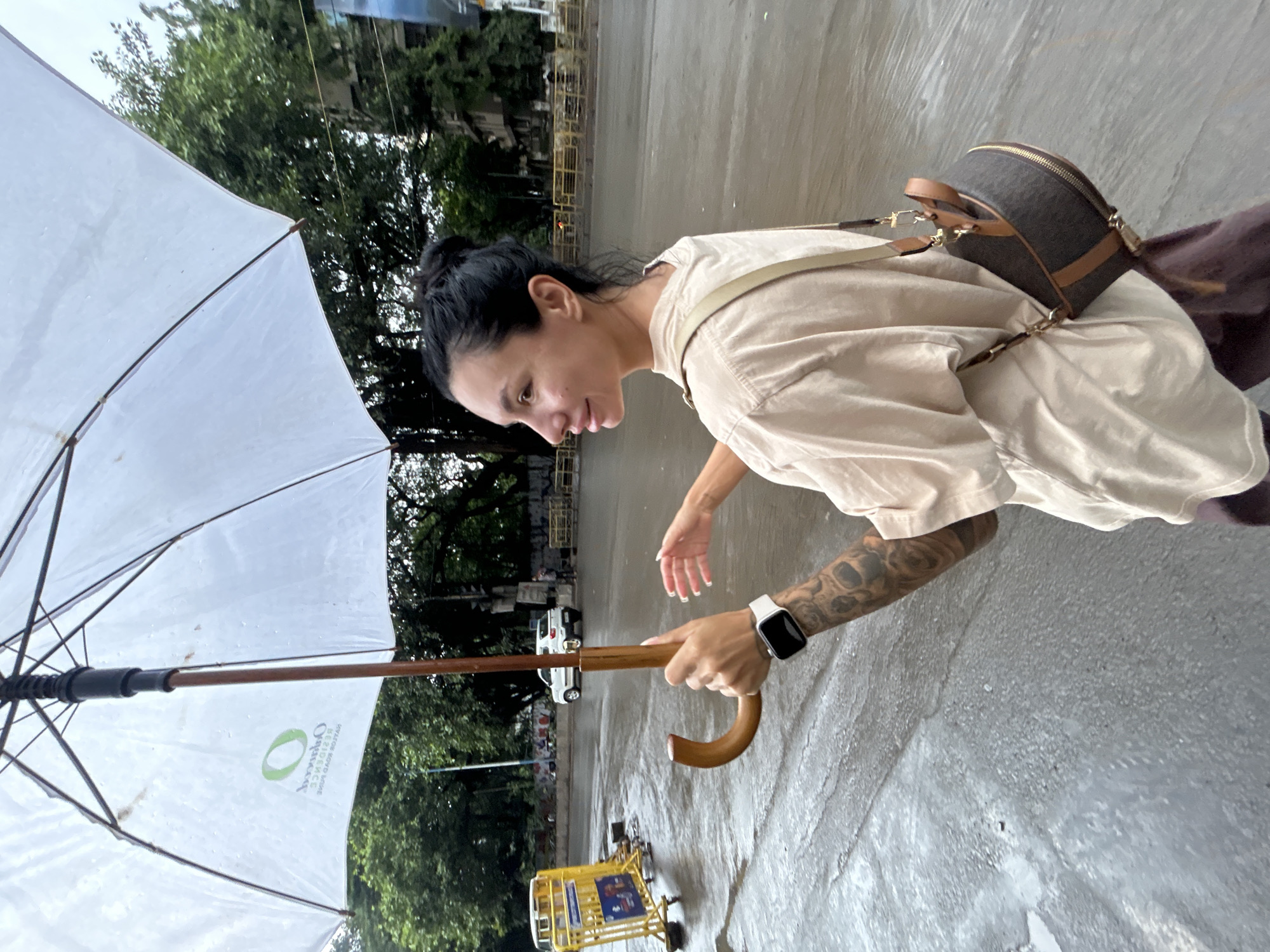 In the city of Pune, India, a woman walks with a white umbrella in the rain. She is wearing a light beige t-shirt and dark purple pants, with a brown and tan patterned crossbody bag. Her hair is pulled back into a bun. On her left wrist, she wears a white smartwatch. The street is wet and reflects the overcast sky. In the background, trees line the street, and a white car is visible. A yellow barricade with blue signage is also present on the left side of the road.