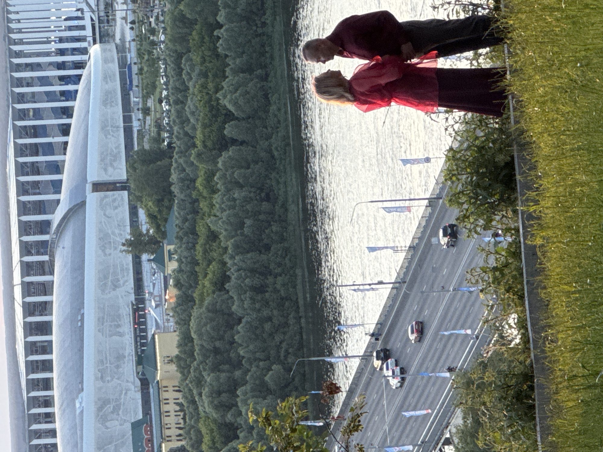 The image shows a wide-angle view of the Nizhny Novgorod Stadium, also known as the Nizhny Novgorod Arena, a modern football stadium with a distinctive curved white roof, situated in Nizhny Novgorod, Russia. In the foreground, a couple stands on a hill overlooking a river and a bridge. The couple, a man and a woman, are facing each other, appearing to be in conversation or sharing an intimate moment. The woman is wearing a long red dress, and the man is in a maroon shirt and dark trousers. The bridge, which carries traffic, spans the river, and several cars are visible on it. The river is calm, reflecting the sky. The background is dominated by the impressive architecture of the stadium and a dense green forest. The overall scene captures a blend of urban infrastructure, natural scenery, and human connection.