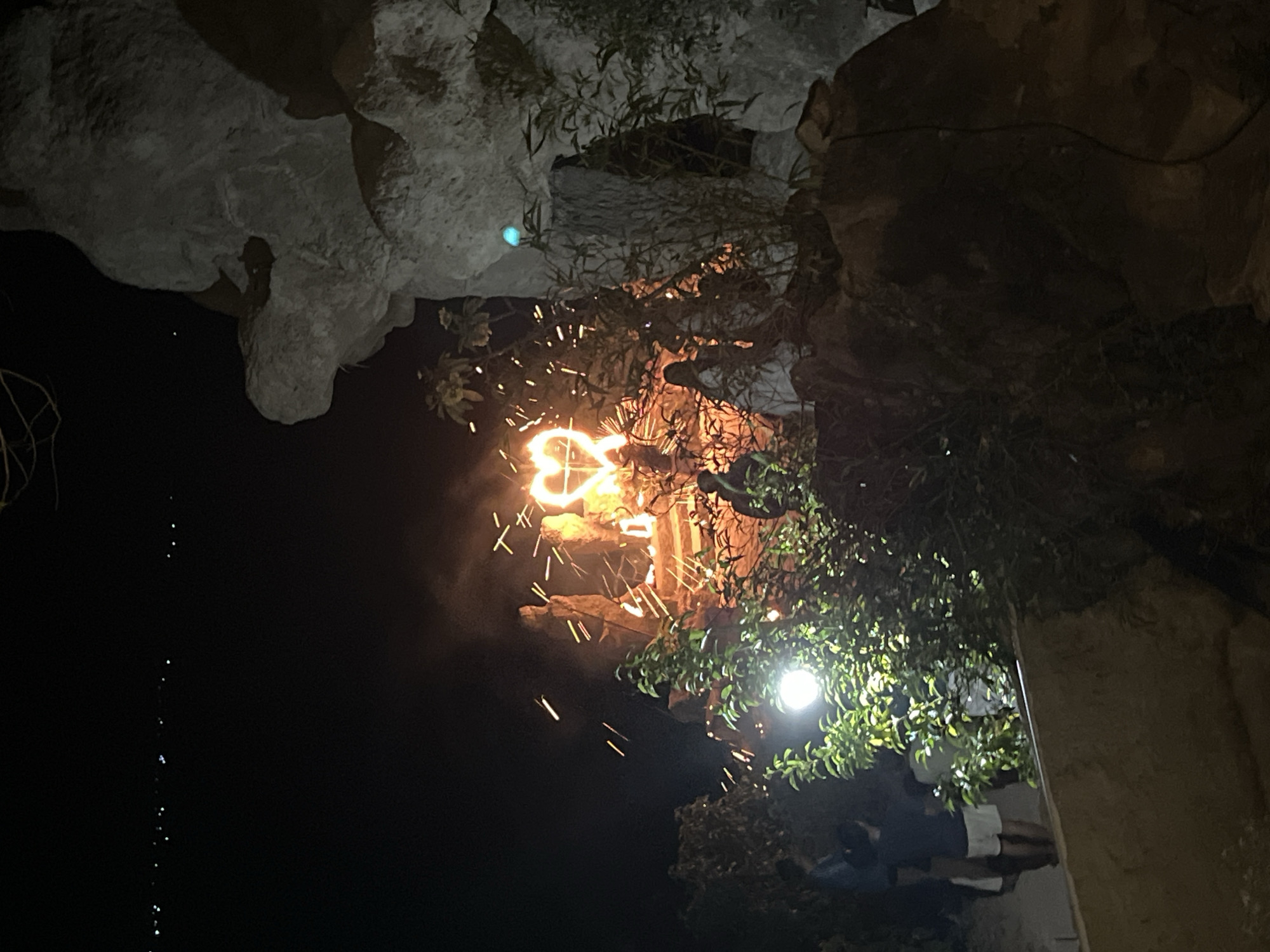 The image depicts a nighttime scene in Kemloko, Indonesia, focusing on a bonfire with a heart shape made of flames. Several individuals are present, appearing to gather around or observe the fire. The environment includes rocky formations and vegetation, with a dimly lit sky in the background dotted with distant lights, possibly from the city or other structures. The overall atmosphere suggests an outdoor event or gathering.