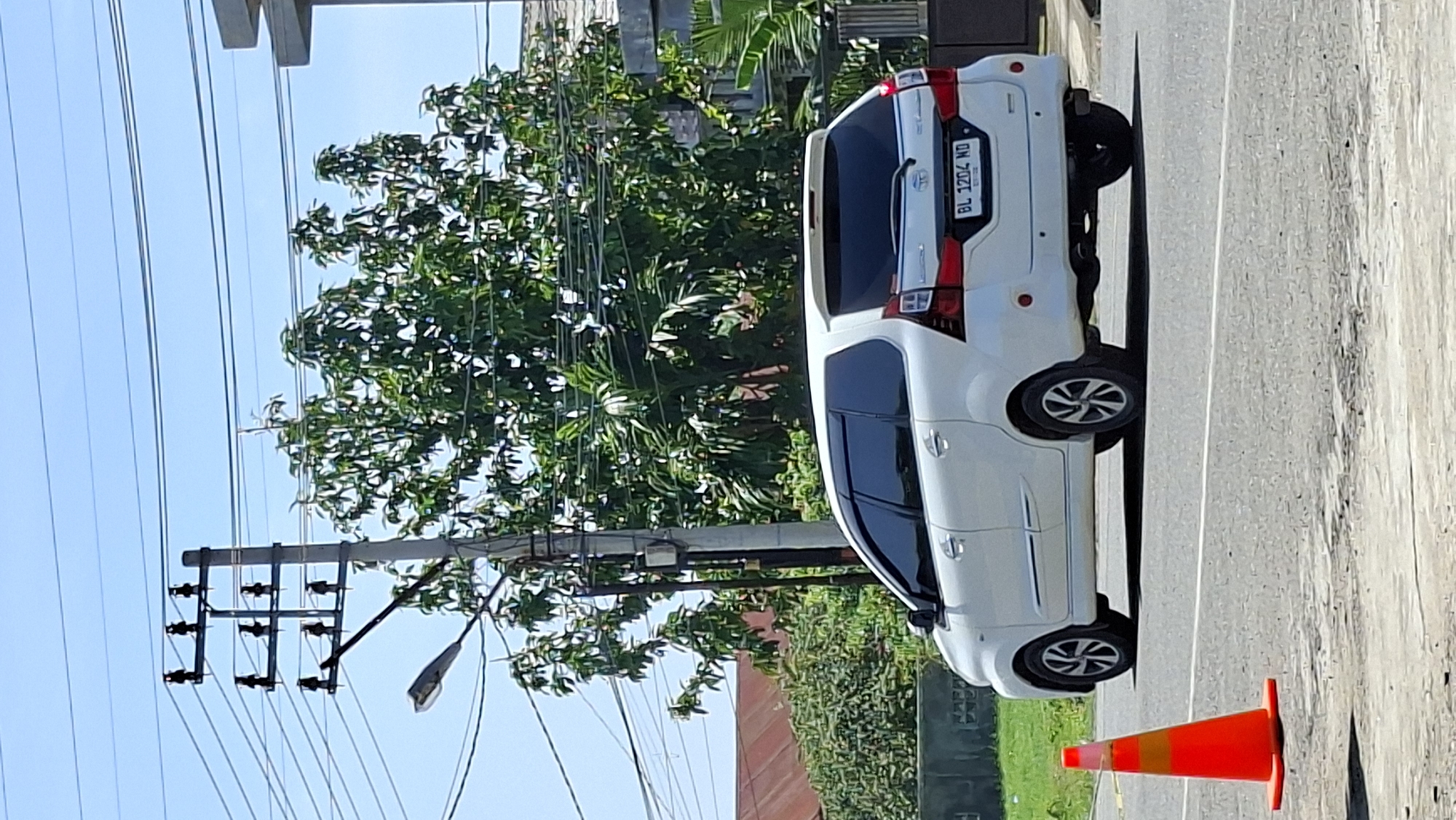 A white minivan, bearing the license plate BL 1204 ND, is traveling on a paved road in Lhokseumawe, Indonesia. The vehicle is viewed from the rear, slightly to the left, and appears to be in motion. In the foreground, an orange traffic cone stands on the right side of the road. Behind the minivan, a lush green tree with dense foliage is visible, obscuring a portion of the sky. Overhead, power lines are strung between utility poles, with one pole positioned to the left of the tree. A street light is also attached to the utility pole on the left. In the background, a reddish-brown roof of a building can be seen, along with other greenery and a portion of a concrete structure on the right. The sky is clear and blue, indicating a sunny day. The scene depicts a typical urban or semi-urban environment with common infrastructure and elements.