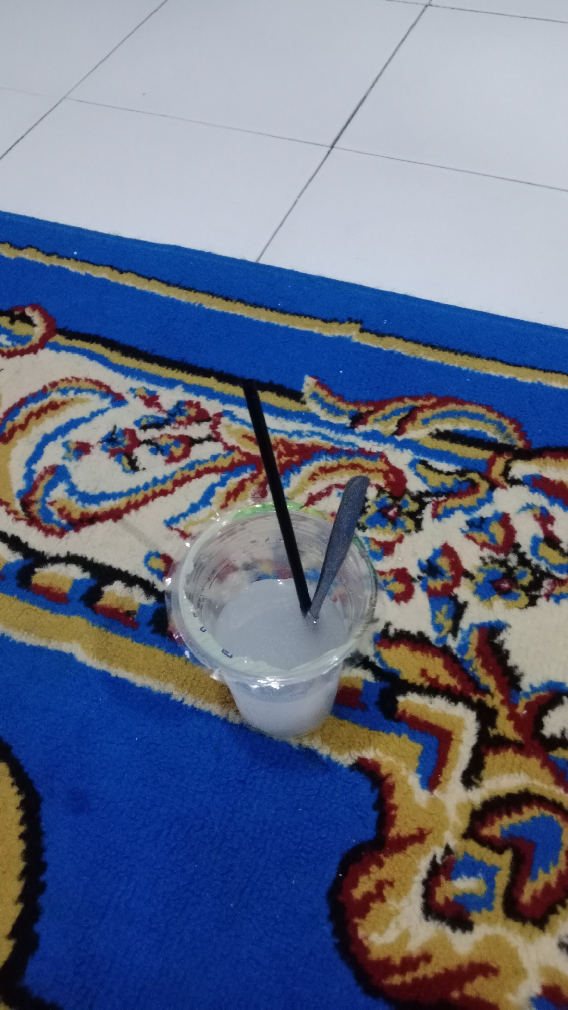 A clear plastic cup filled with a white, opaque liquid sits on a blue, red, and yellow patterned rug. Two dark-colored utensils, a straw and a spoon, are submerged in the liquid. The rug is placed on a floor tiled with white square tiles. The scene appears to be indoors, possibly in a domestic setting. There are no people visible in the image. The lighting is dim, suggesting an indoor environment without direct sunlight.