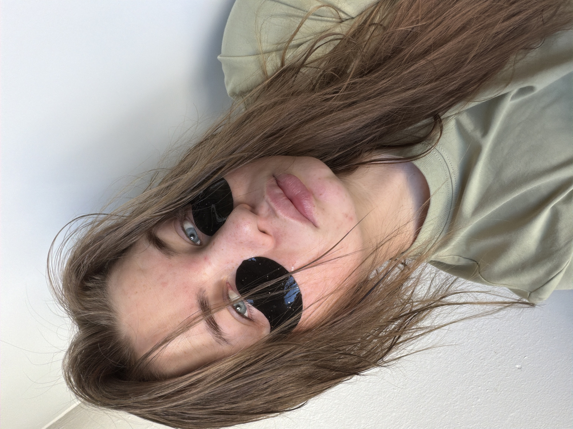 A woman with light brown hair and blue eyes is wearing round black sunglasses. She is wearing a light green shirt. The location appears to be indoors with a white wall in the background. It is not possible to determine if this image was taken in Chaikovsky, Russia or any other city. There is not enough information available to conclude that the location is in the city of Chaikovsky, Russia.