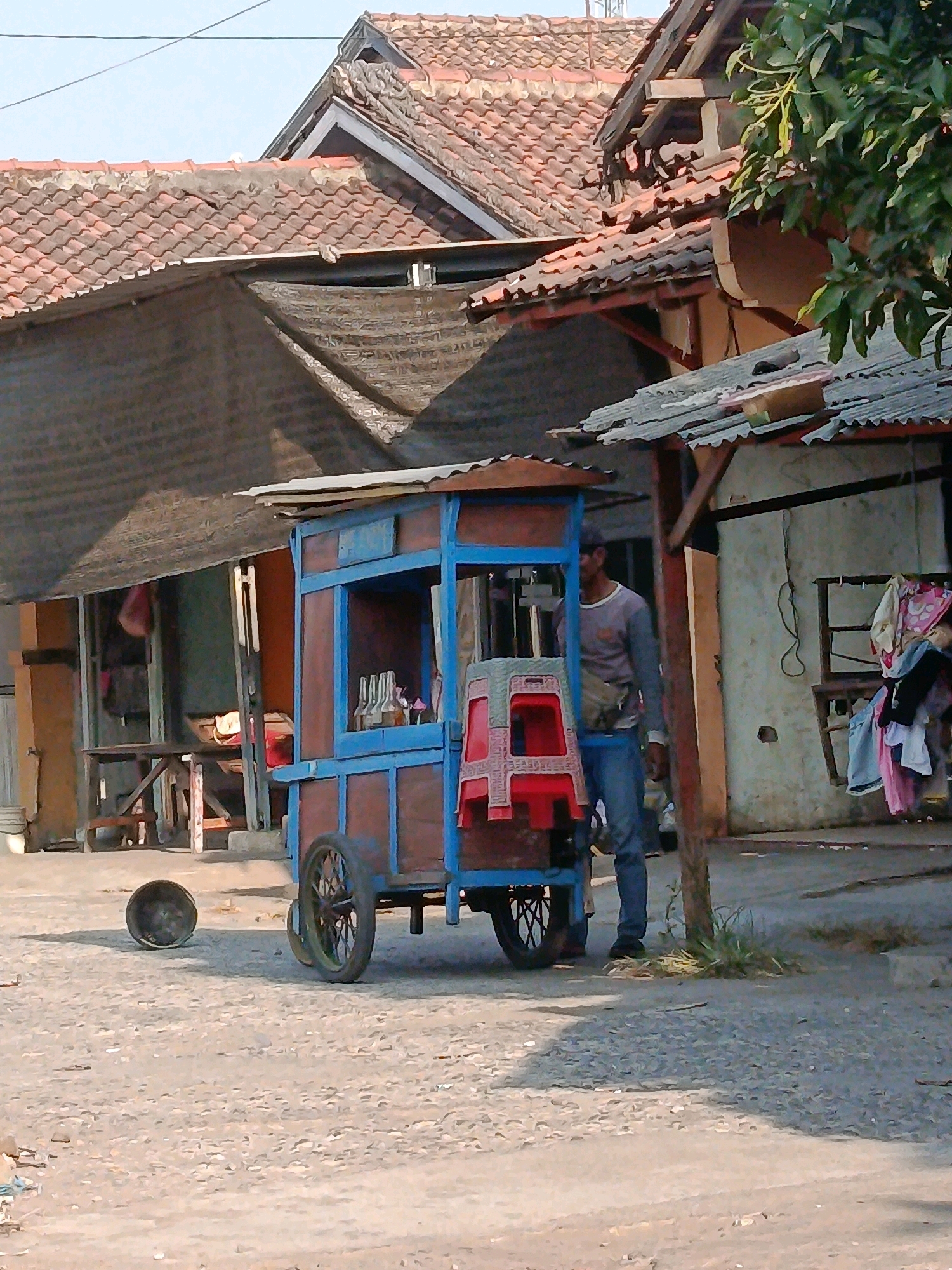 A man stands beside a small, wheeled cart in a dusty, outdoor setting. The cart, made of wood and painted blue and brown, has a corrugated metal roof and displays several bottles on shelves. Two red plastic chairs are stacked on the side of the cart. The man, wearing a grey long-sleeved shirt, blue jeans, and a sling bag across his chest, appears to be tending to the cart. In the foreground, a dark, round object, possibly a bucket or a pot, rests on the ground. Behind the cart, a building with a tiled roof and open storefronts is visible, with laundry hanging on a rack outside one of the structures. The overall environment suggests a street scene in a residential or commercial area. The location is identified as Jobokuto, Indonesia.