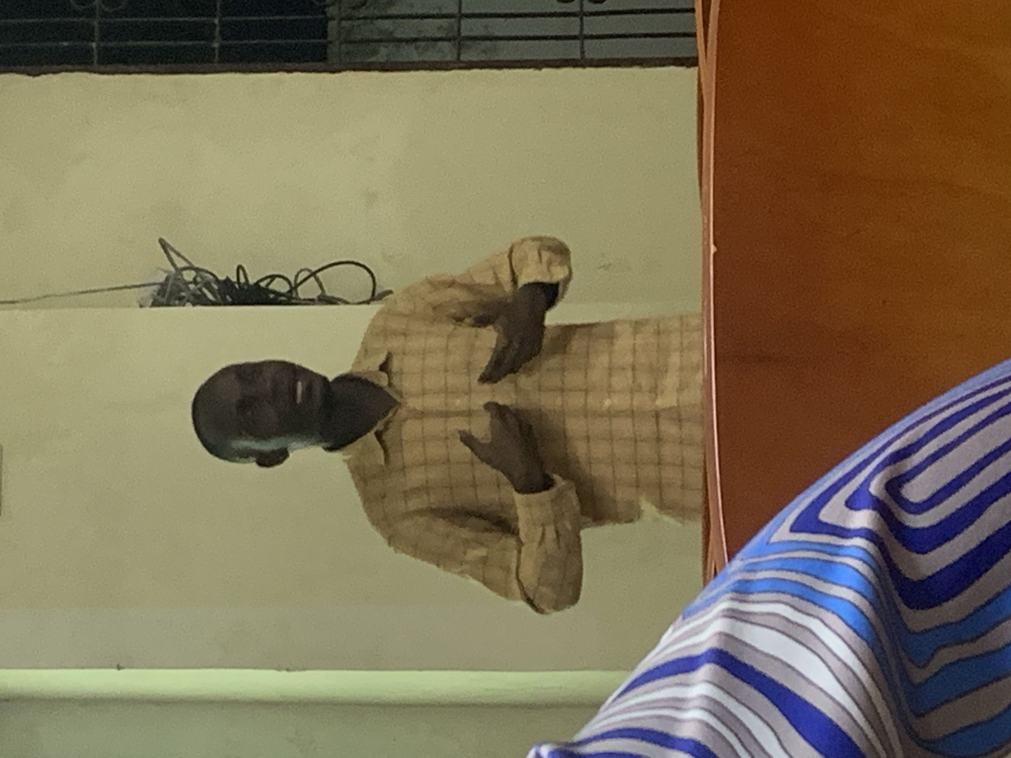 A person with dark skin is wearing a checkered shirt. Their hands are raised, with fingers slightly bent. The person is positioned against a light-colored wall. In the background, there is a black wire and window bars. In the foreground, part of a blue and white striped material is visible. The location could be Dar es-Salaam, Tanzania. The exact location within the city is not clear.