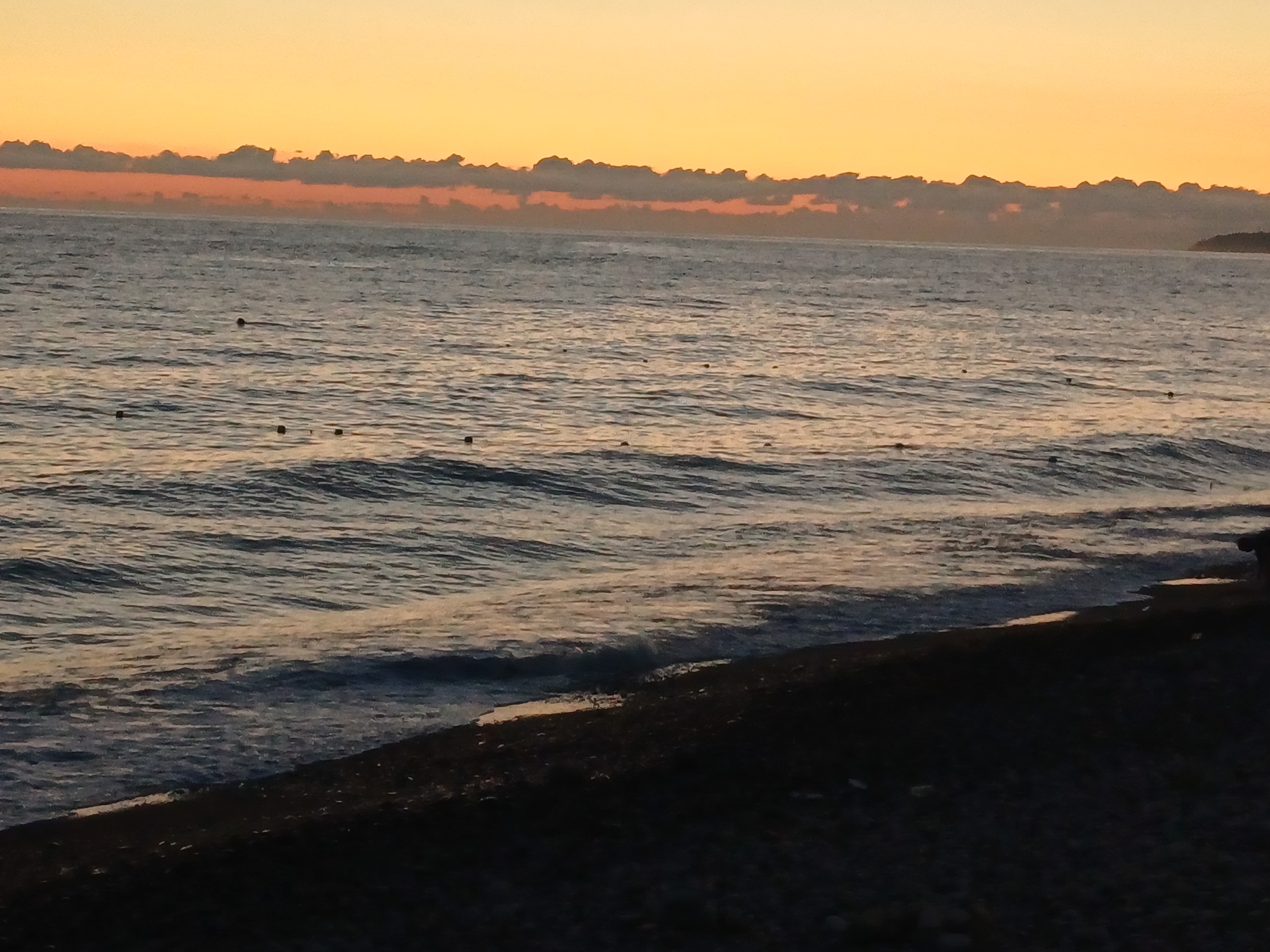 The image depicts a serene beach scene at sunset in Sochi, Russia. The sky is a gradient of warm orange and yellow hues, transitioning to a soft blue at the zenith. A band of dark, fluffy clouds stretches across the horizon, silhouetted against the vibrant sky. The sea is calm, with gentle waves lapping the shore. Small buoys or markers are scattered across the water's surface. The foreground is a dark, pebbled beach, sparsely populated and seemingly undisturbed. The overall atmosphere is tranquil and peaceful, characteristic of a sunset by the Black Sea.