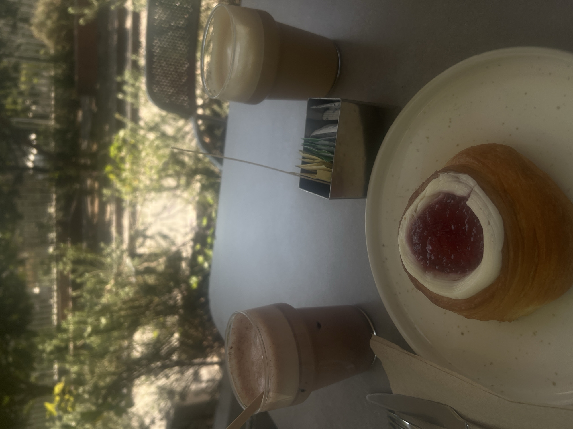 The image depicts a breakfast or brunch setting, likely outdoors. On a table, there's a croissant filled with red jam and topped with a cream-like substance, presented on a speckled white plate. Beside the plate, there are two glass cups containing beverages, one with a lighter, milky coffee color and another with a darker, mocha-like hue. There is a metal sugar packet container between the plate and the cup of lighter coffee. Cutlery is visible near the bottom of the frame. The background features a blurred view of greenery, suggesting a patio or garden setting. Based on the food and general setting, it is possible this photograph was taken in Mexico City, Mexico.