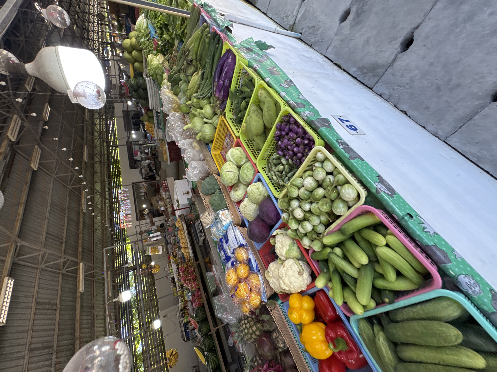 The image shows an outdoor market stall filled with a variety of fresh produce, predominantly vegetables. The stall is set up along a concrete wall, with a corrugated metal roof structure overhead, suggesting an open-air market or covered walkway.
The main subjects are the vegetables, neatly arranged in colorful plastic baskets. We can see cucumbers in both pink and light blue baskets, as well as small green eggplants in a light green basket. Other vegetables include purple eggplants, cauliflowers, and what appear to be gourds or squashes. Further down the stall, red and yellow bell peppers are visible, along with other fruits and vegetables that are less distinct. There are also some items wrapped in plastic bags, possibly for sale.
In the background, there are glimpses of what looks like the interior of a market or building, with metal beams and lighting fixtures. Decorative lights and what appear to be large, ornamental lamps hang from the ceiling structure. Some bags of produce are also visible in the background, suggesting a busy market environment.
The scene is brightly lit, possibly indicating daytime. The overall impression is one of abundance and freshness. There is a price tag with the number "67." visible on one of the green decorative borders of the stall. No people are clearly visible in the frame, but the presence of a market stall implies human activity. The concrete wall in the background has several circular holes, possibly drainage or ventilation features.