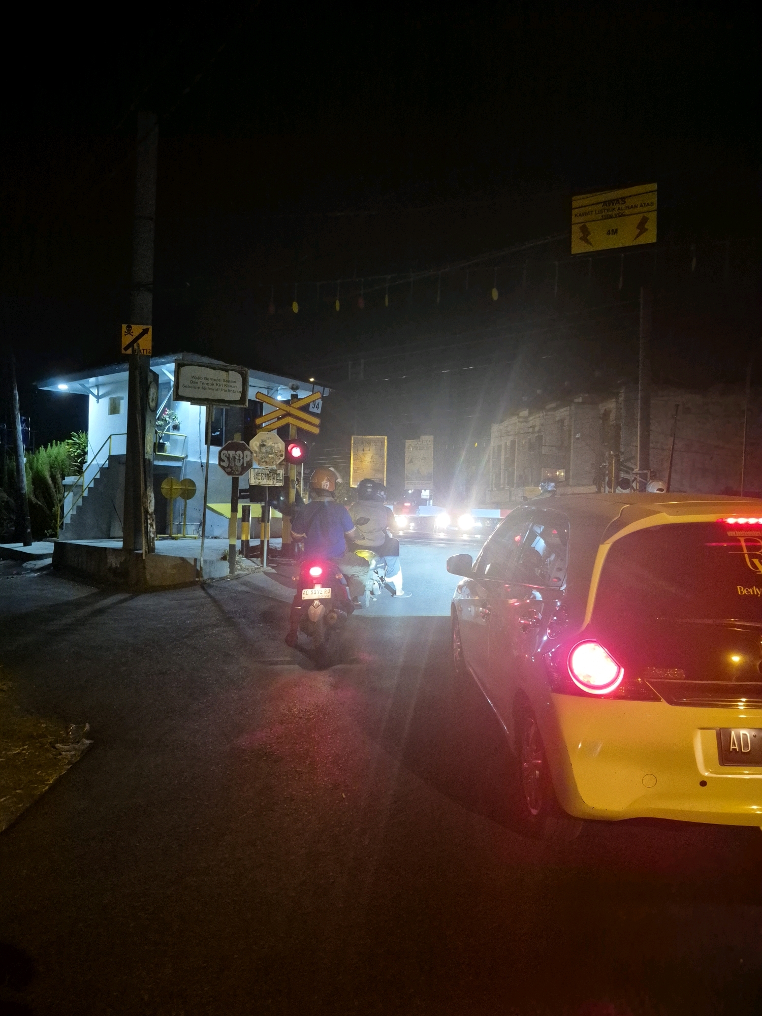 The image depicts a nighttime scene at a railroad crossing in Surakarta, Indonesia, likely near a residential area given the presence of trees and a small building. In the foreground, a yellow car with its brake lights illuminated is stopped. Behind it, two motorcycles are also stopped, with the rider of the first motorcycle visible from the back, wearing a blue shirt and a helmet. The second motorcycle has a rider with a helmet, seen from the side.
To the left, a small, white, elevated building with stairs and a blue-lit entrance stands near the railroad tracks. A traffic signal showing a red light is visible, along with various signs. One sign, partially obscured, appears to be a "STOP" sign. Other signs include railway crossing signals with yellow and white stripes. A warning sign with a skull and crossbones and the text "AT12" is mounted on a pole. Further back, streetlights and the headlights of other vehicles create a hazy glow. Overhead, a yellow warning sign with lightning bolt symbols and the text "4M" suggests a clearance limit, possibly for electrical wires. The road surface is asphalt, wet in some areas, reflecting the ambient light. The overall atmosphere is one of waiting, common at a train crossing with a red light.