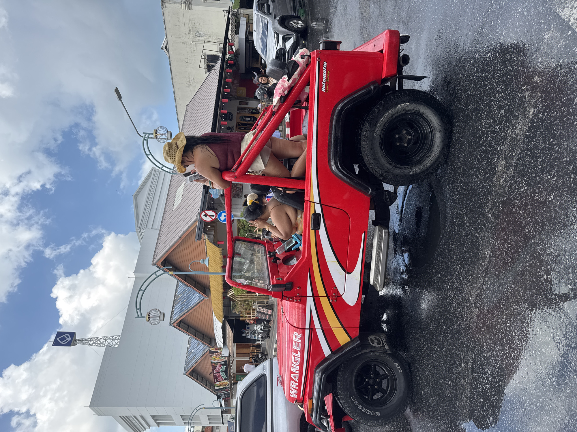 The image captures a red Jeep Wrangler, heavily modified with a roll cage and exterior graphics, parked on a wet city street in Phuket City Municipality, Thailand. The pavement reflects the sky and surrounding buildings, suggesting recent rain. Inside the Jeep, two women are visible; one is wearing a straw hat and looking at her phone, while the other is seated in the front. The street scene includes buildings with awnings and various shops. Other vehicles are also parked nearby, and streetlights are present. The Wrangler appears to be the central subject, contrasting with its urban surroundings.