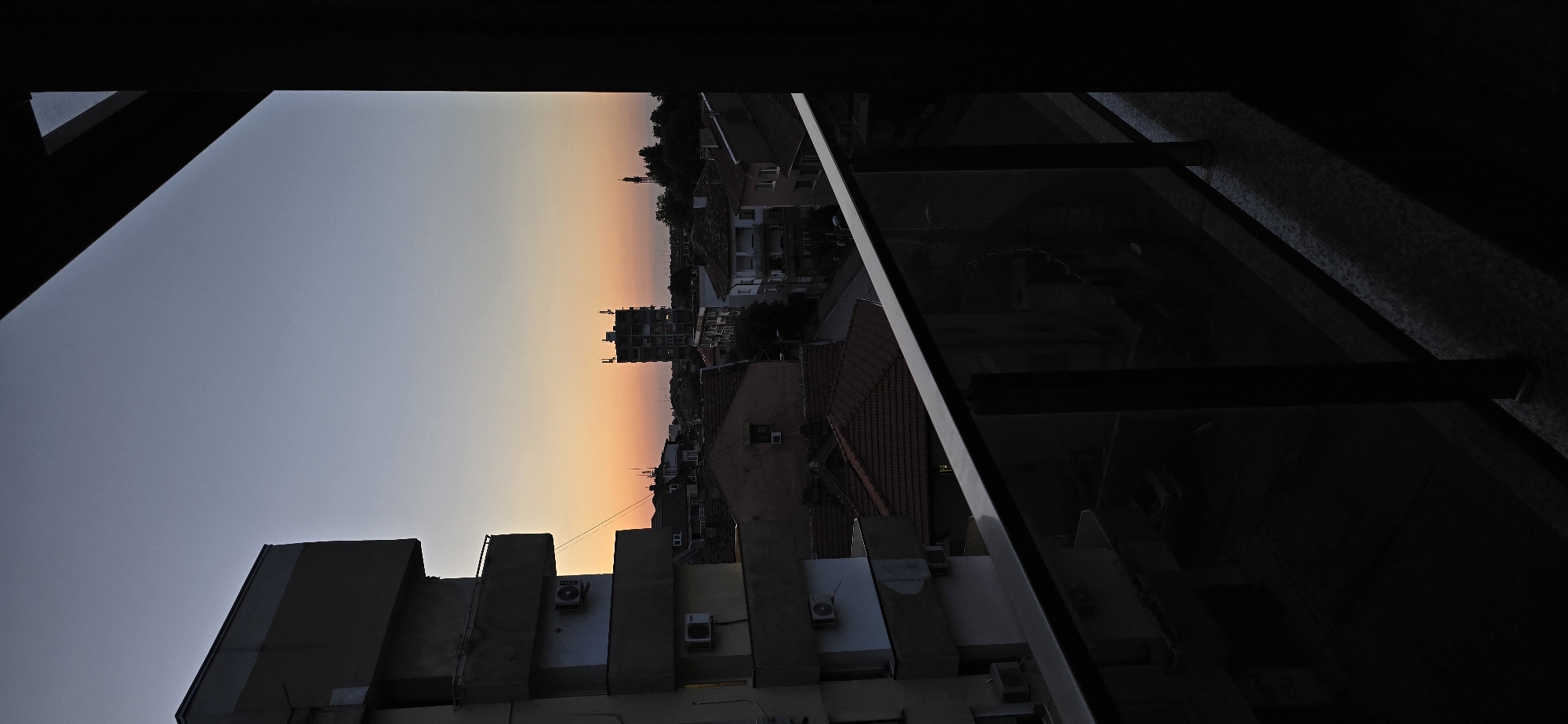 The image presents a forensic view of an urban landscape at twilight, observed from a balcony. The sky transitions from a warm orange and pink glow at the horizon, indicative of either sunset or sunrise, to a cool, muted blue-grey towards the zenith. No clouds are visible.
In the immediate foreground, a dark balcony railing, featuring a metallic top bar and vertical supports with a transparent or dark glass panel, extends across the lower third of the frame. The railing and the balcony floor are rendered in deep shadow, almost entirely silhouetted.
To the left, a multi-story apartment building, characterized by an architectural style suggestive of the mid-20th century with protruding rectangular balconies and multiple external air conditioning units mounted on its facade, stands in prominent silhouette. Its surfaces appear dark grey and shadowed.
The midground reveals a dense cluster of buildings, predominantly residential. Many feature distinct reddish-brown tiled roofs. Interspersed among these are buildings with lighter, weathered facades, some showing signs of disrepair. A taller, slender building with structures on its rooftop stands out towards the center-right, also in silhouette. A narrow street or alley is visible winding through the buildings. Sparse trees and foliage are present, particularly on the right, contributing to the shadowed landscape. A faint, slender spire or antenna is barely discernible on the distant horizon line to the right.
The overall illumination is low, with the ambient light originating from the horizon. No individuals are visible, and no specific actions are depicted. The scene is static and devoid of human presence. The location is an unknown city or town in an unknown country.