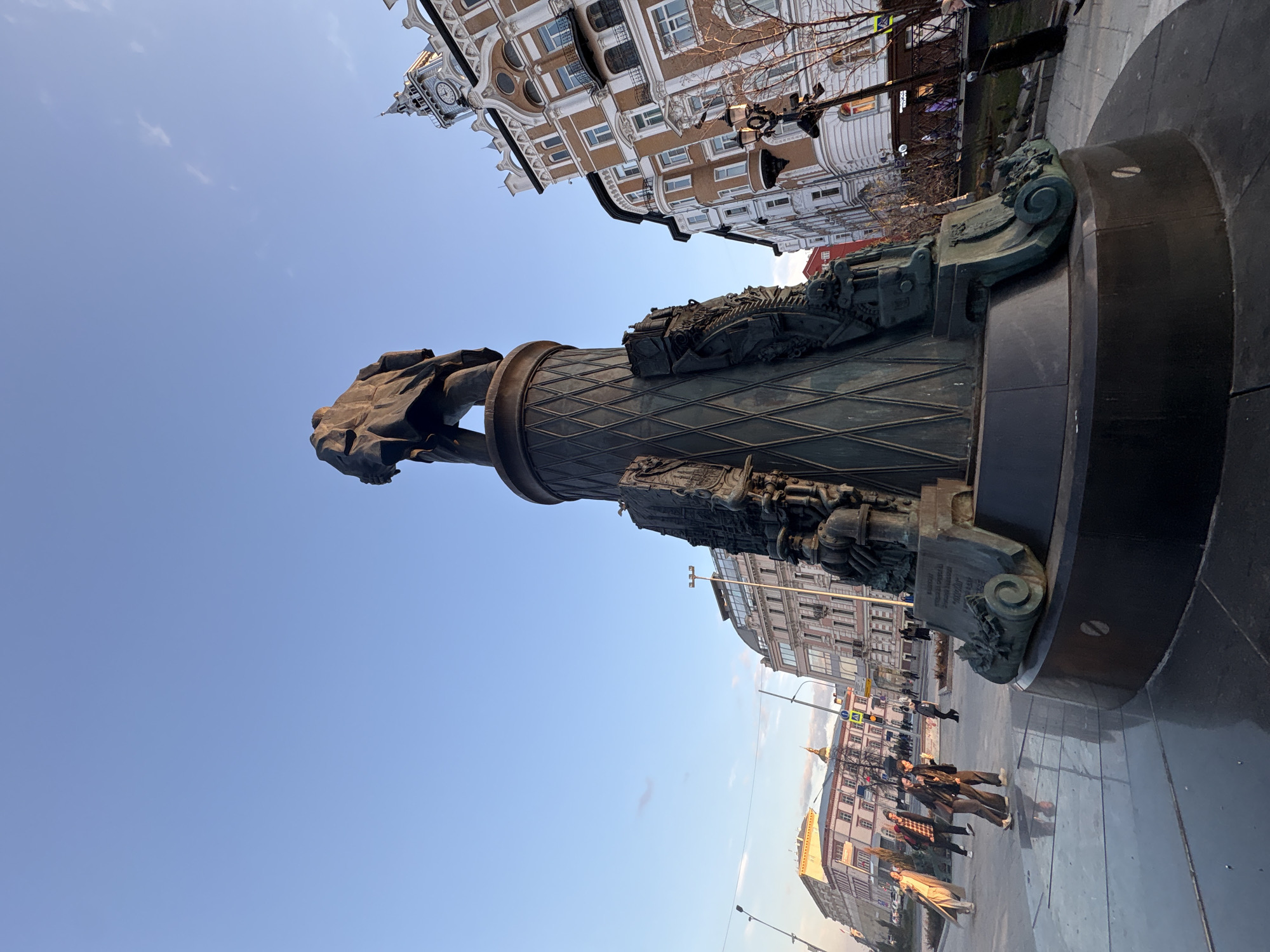 The image depicts a monument in Moscow, Russia, against a backdrop of buildings and a blue sky. The monument features a statue of a person atop a decorated pedestal. The pedestal exhibits detailed carvings and ornamentation. Several people are walking on the pavement around the monument. Buildings with intricate architectural designs are visible in the background. The sky is clear, with minimal cloud cover.