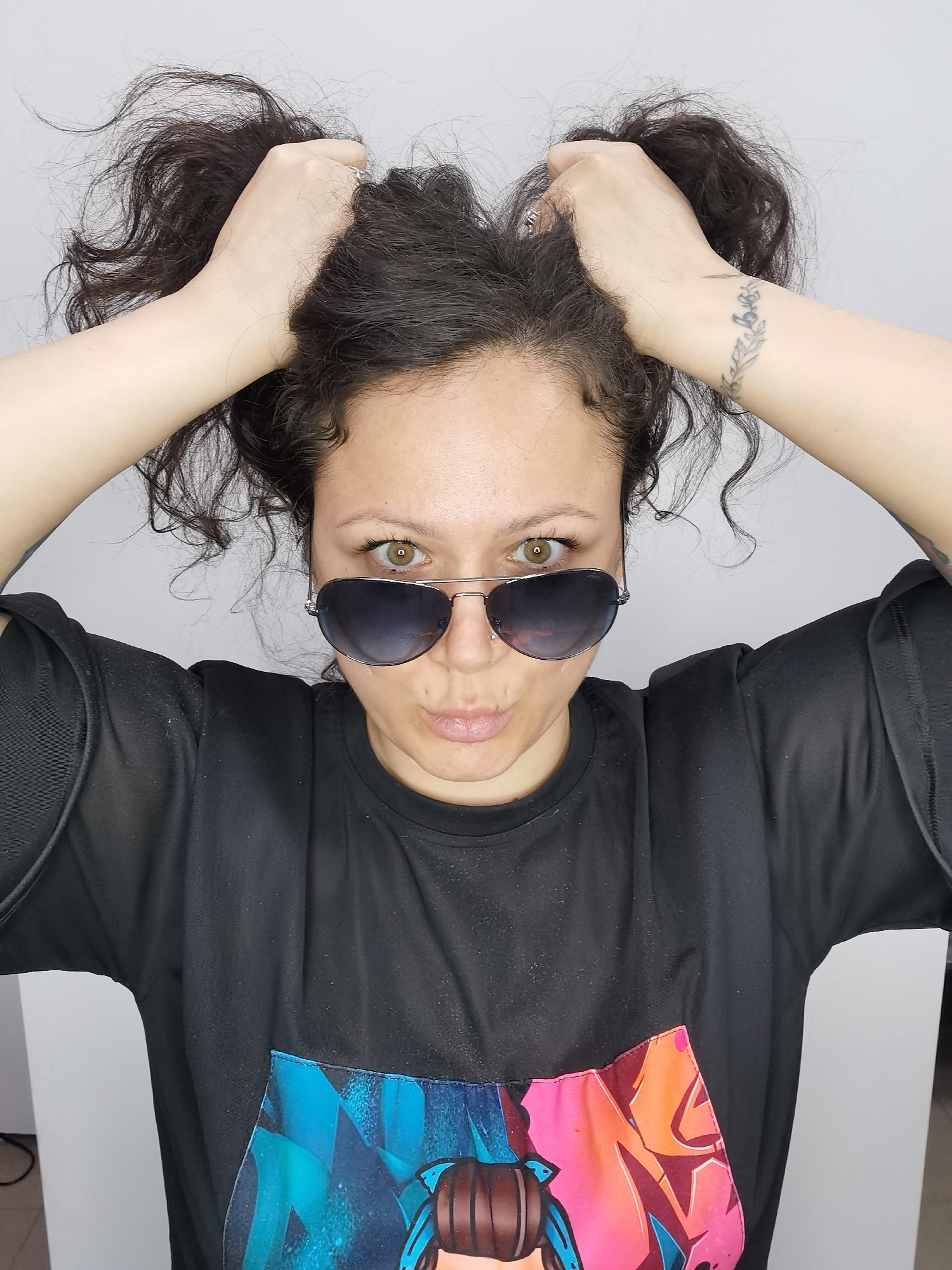 A woman poses indoors. She has dark, curly hair pulled up into two messy buns with her hands. She is wearing aviator sunglasses, a black t-shirt with a colorful graphic design, and has a tattoo on her left wrist. The background is a plain, light-colored wall. There is no evidence suggesting the location is in Chaikovsky, Russia.