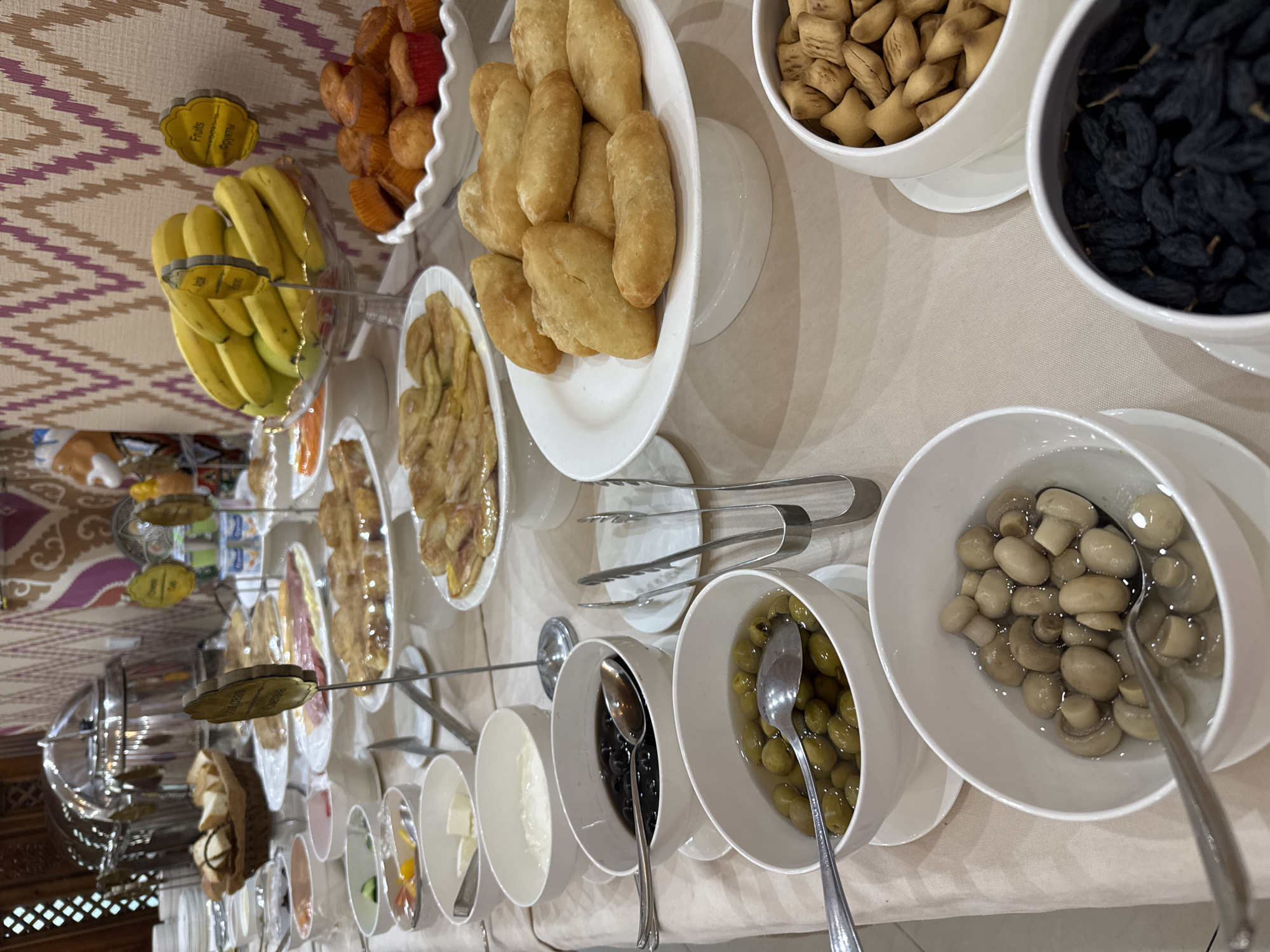 The image depicts a breakfast buffet in Samarkand City, Uzbekistan. On a white tablecloth, there are various food items arranged in white bowls and plates. These include fruits (bananas), pastries, small star-shaped crackers, raisins, pickled mushrooms, olives, and a selection of condiments and bread. There are also serving utensils like spoons and tongs near the food items. The background is somewhat blurred, indicating a hotel or restaurant setting.