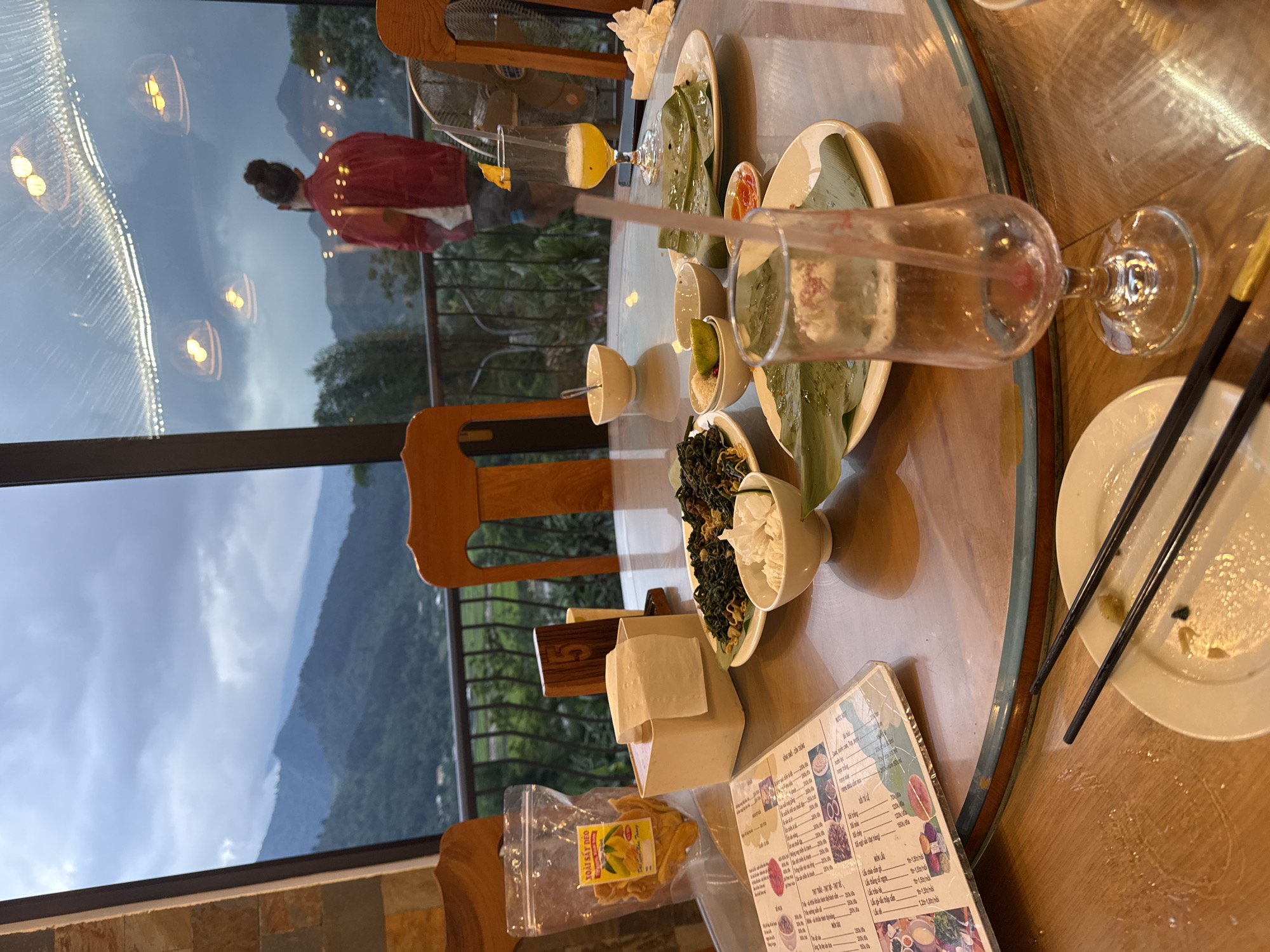 The photograph captures a dining scene in Tú Lệ, Vietnam, positioned near a window. A round wooden table dominates the foreground, laden with dishes and remnants of a meal. There are plates with chopsticks, a glass with a straw, and a bowl containing a salad. A menu and packaged snacks are also visible. Outside the window, a landscape of mountains and valleys is visible, suggesting a high-altitude or elevated location. Reflected in the glass is the silhouette of a person in a red jacket, further emphasizing the interior-exterior dynamic of the shot.