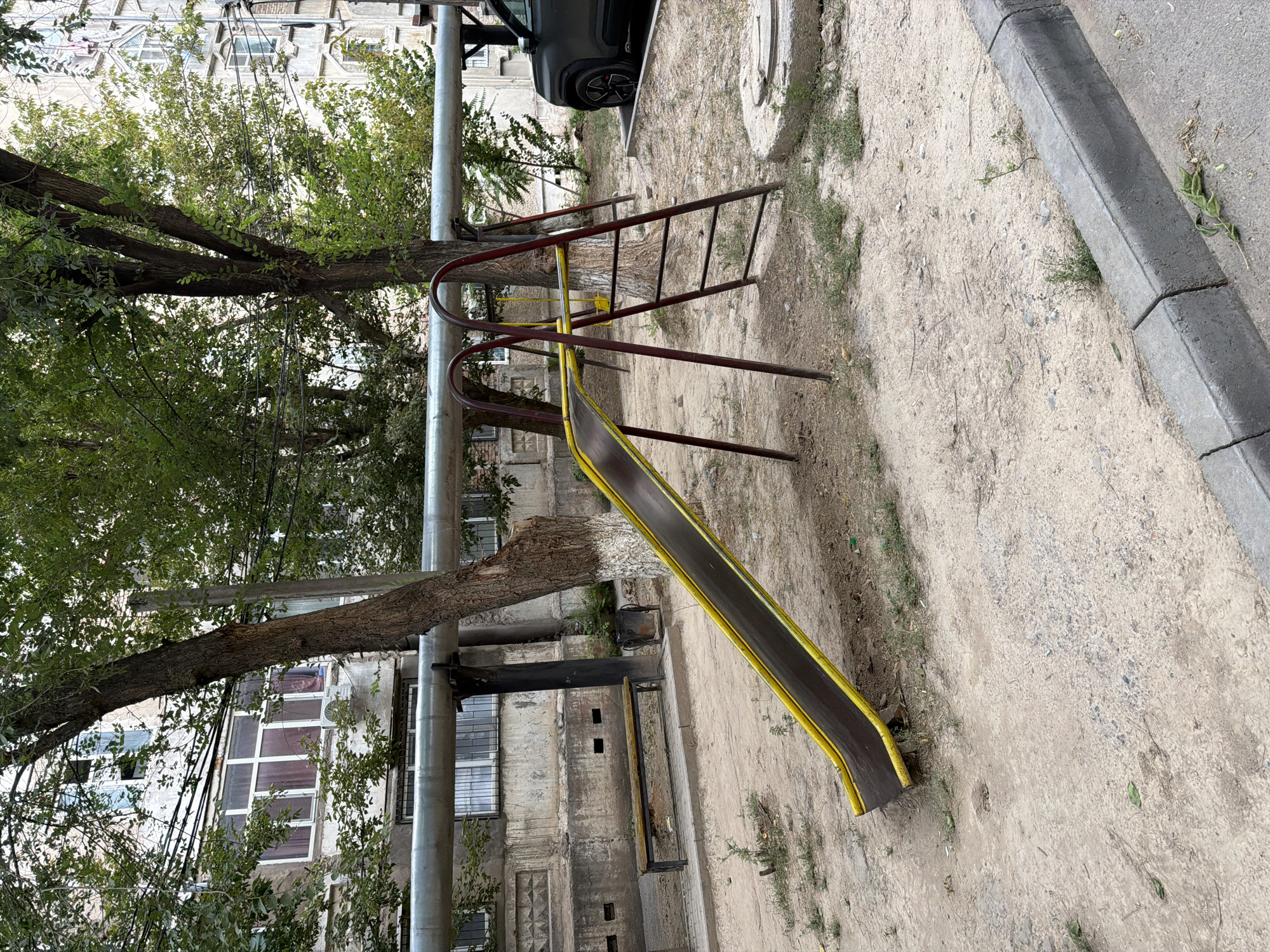 The image depicts an outdoor scene in Shymkent, Kazakhstan, featuring a children's playground slide set against a backdrop of an apartment building and trees. The slide itself is made of dark material with yellow trim and is supported by a red metal A-frame structure with steps. To the left of the slide, a simple wooden bench with a metal frame is visible, alongside a trash can and a large pipe running horizontally across the frame. The ground is covered in sand and dirt, with scattered patches of grass and small plants. In the background, a multi-story apartment building with numerous windows can be seen. A dark-colored SUV is parked to the right of the frame. The overall environment suggests an urban residential area with limited public amenities.