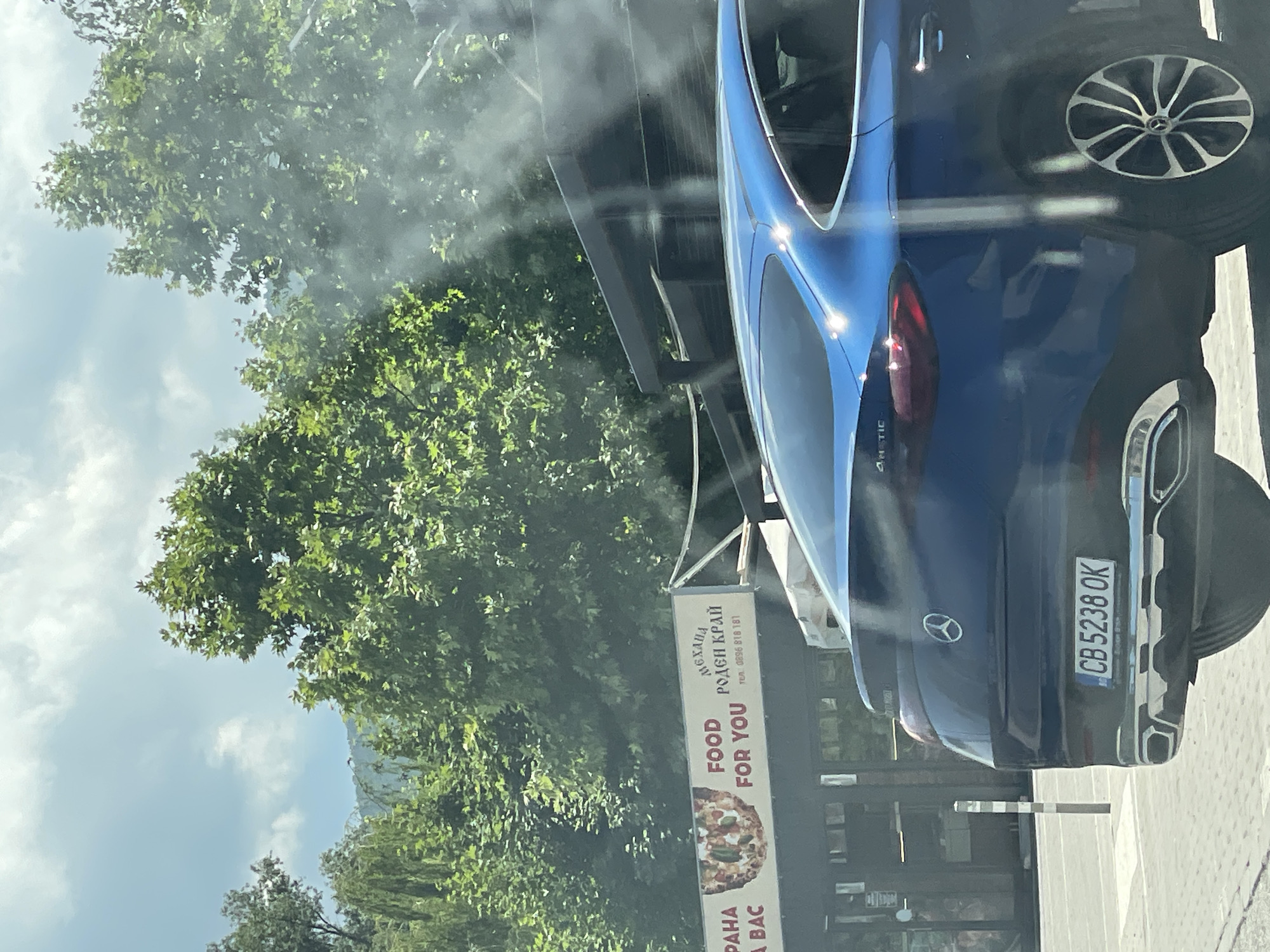 An outdoor, eye-level medium shot captures a blue Mercedes-Benz parked behind what appears to be a food establishment. The Mercedes-Benz is facing left, with its rear bumper and taillights prominently displayed. The license plate, "CB 5238 OK," is clearly visible. To the left of the car, a sign for a restaurant reads "FOOD FOR YOU" and "МЕХАНА РОДЕН КРАЙ" in Cyrillic, along with a phone number. The background consists of lush green trees and a cloudy sky. The overall scene suggests a daytime setting in a city.